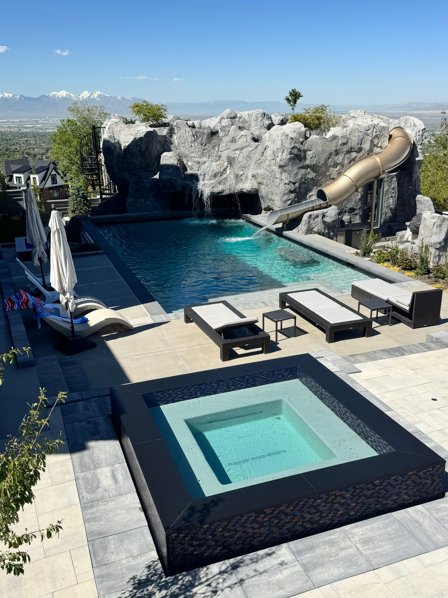 An elevated outdoor pool and square hot tub with a rock feature and slide against a backdrop of snow-capped mountains.