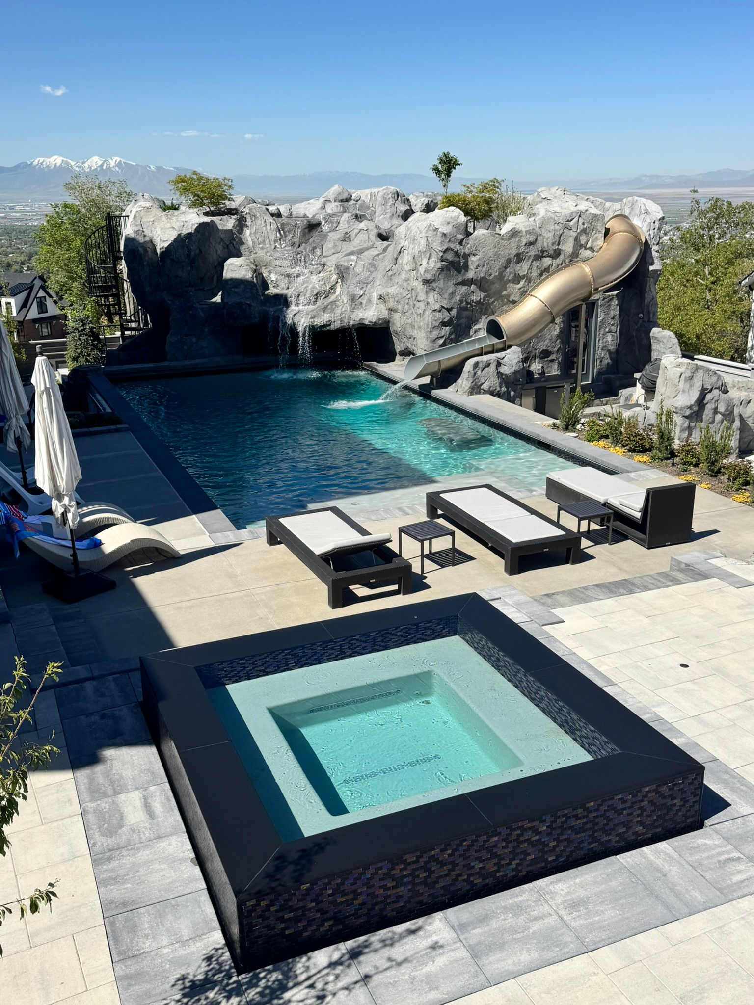 A high-angle view of a backyard pool area with a square hot tub, swimming pool, water slide, and lounge chairs.