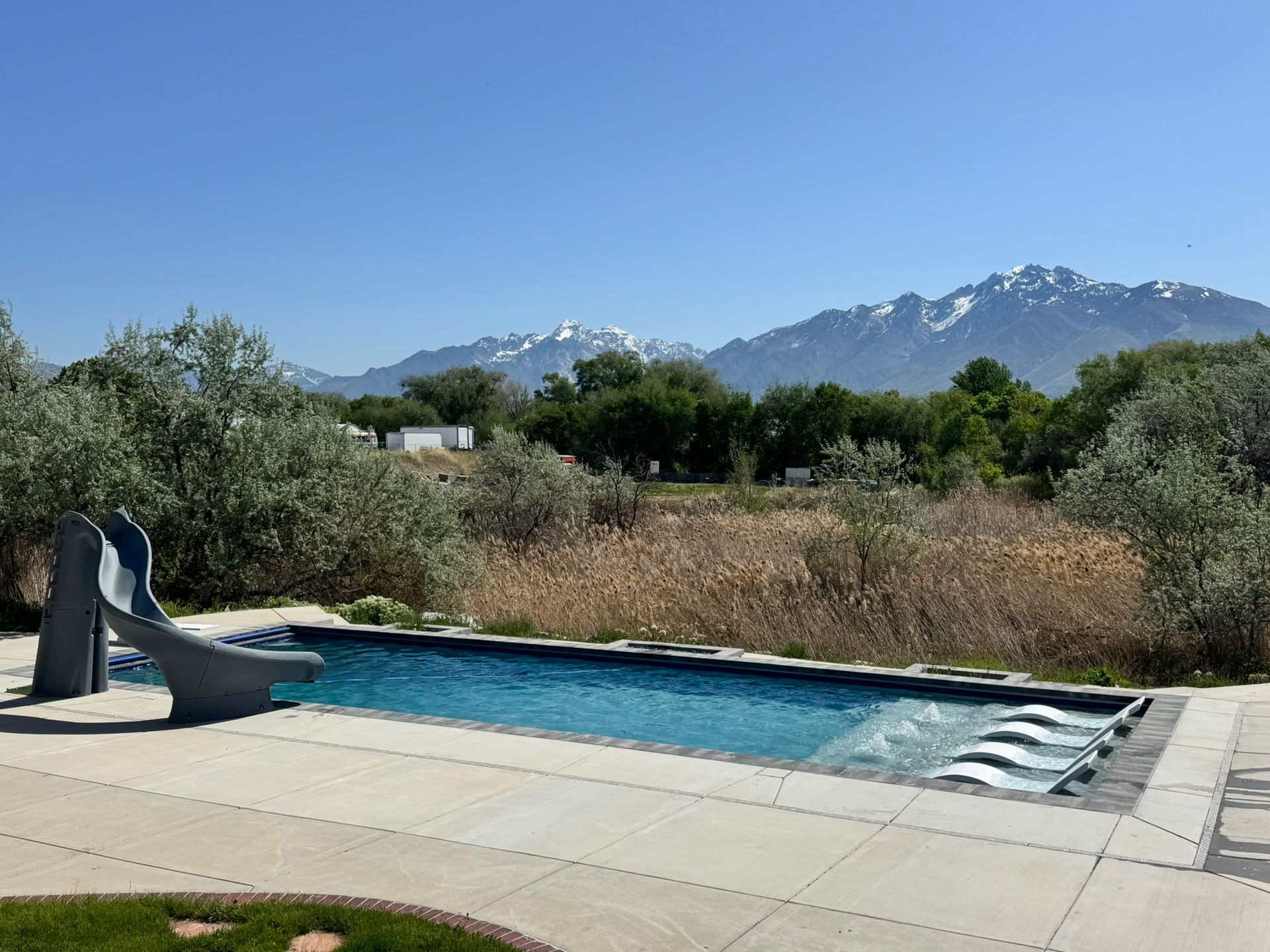 A backyard swimming pool with a slide and bubblers, set against a backdrop of snow-capped mountains and green trees.