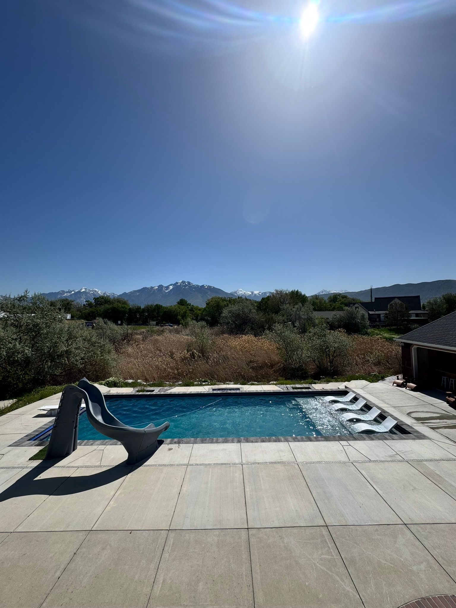 A swimming pool with a slide, surrounded by a concrete patio, featuring mountain views under a bright, sunny sky.