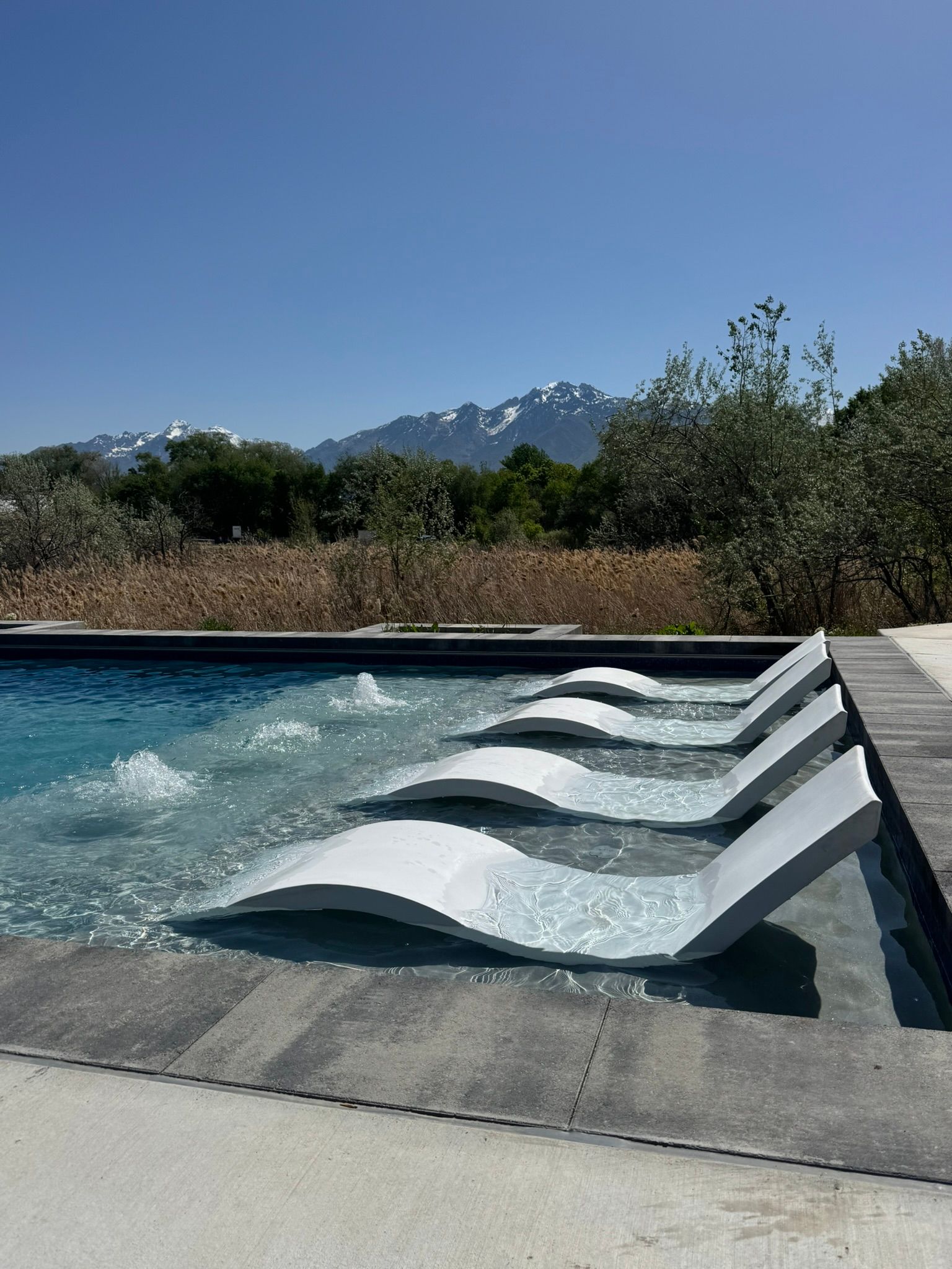 White lounge chairs sit partially submerged in a pool shelf against a backdrop of mountains and trees under a blue sky.