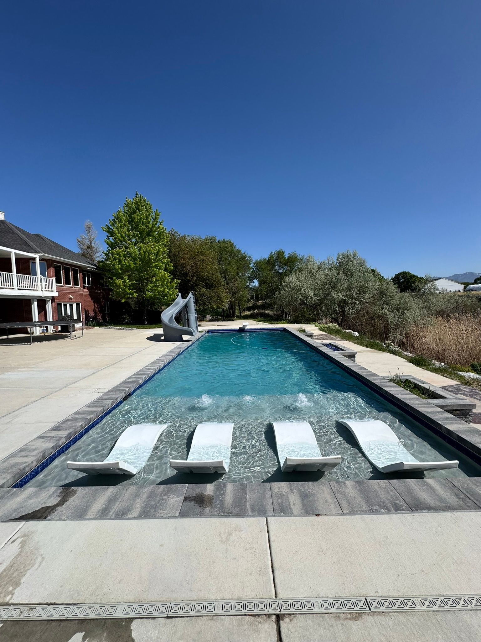 A rectangular swimming pool under a bright blue sky, with four white lounge chairs partially submerged in the shallow end.