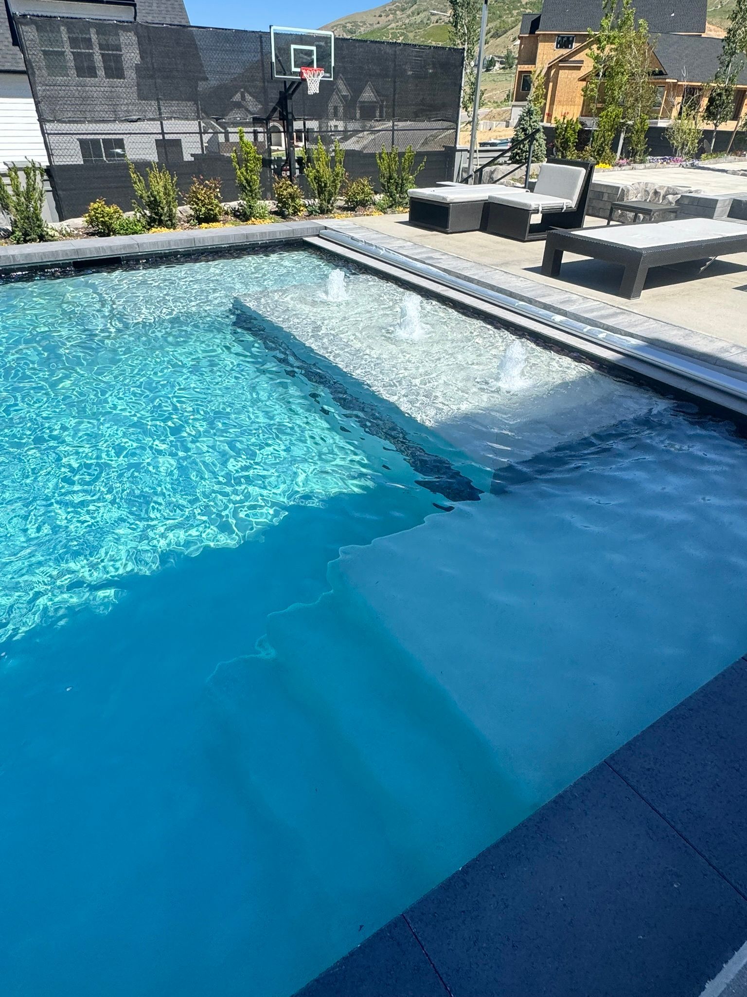 A blue rectangular swimming pool with a shallow tanning ledge, water bubblers, and stairs, set in a sunny backyard.