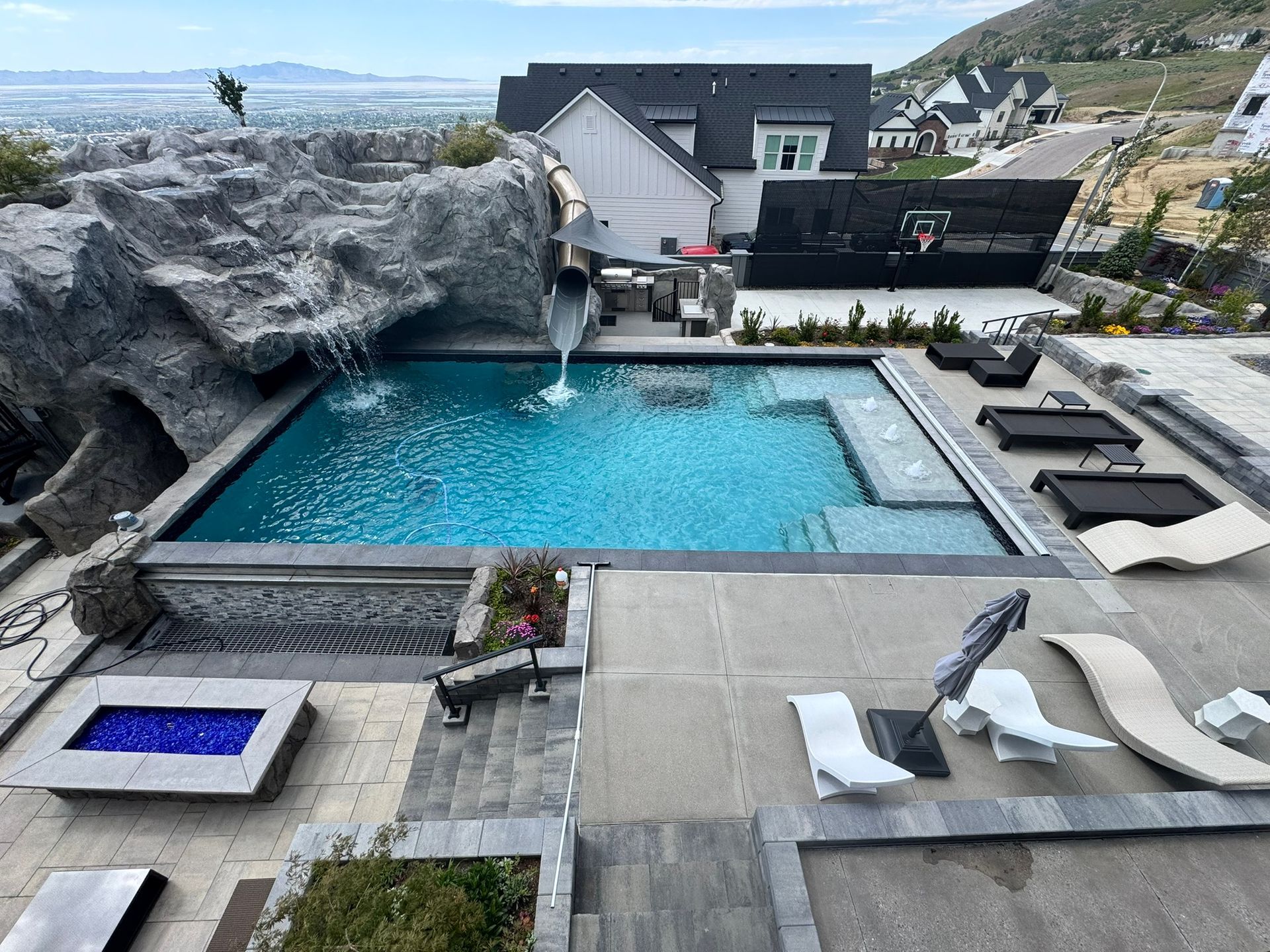 A backyard swimming pool with a rock waterfall, slide, lounging area, and fire pit overlooking a mountain landscape.