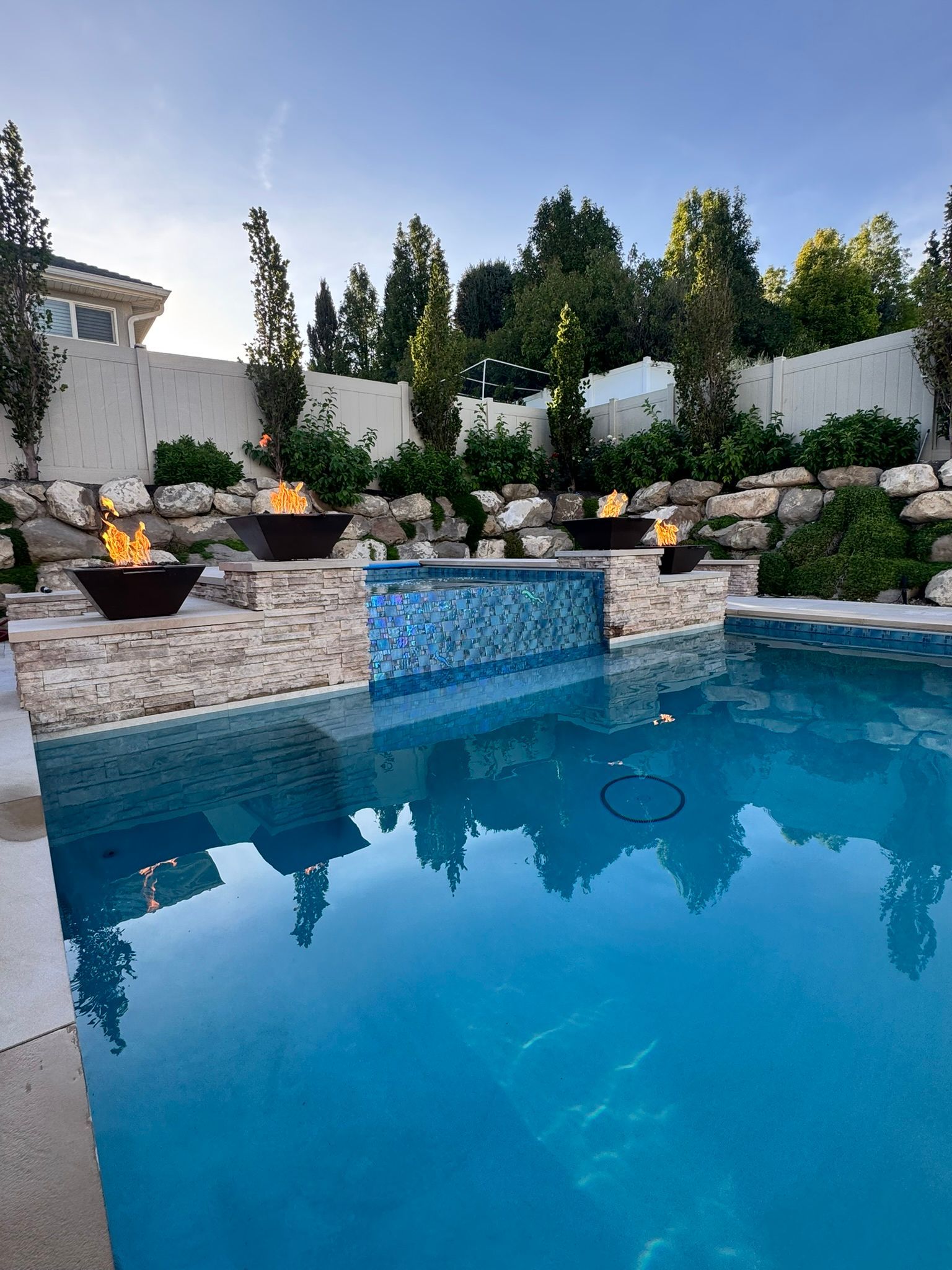 A swimming pool with a stone wall feature, a blue tile water feature, and three lit fire bowls on a sunny day.