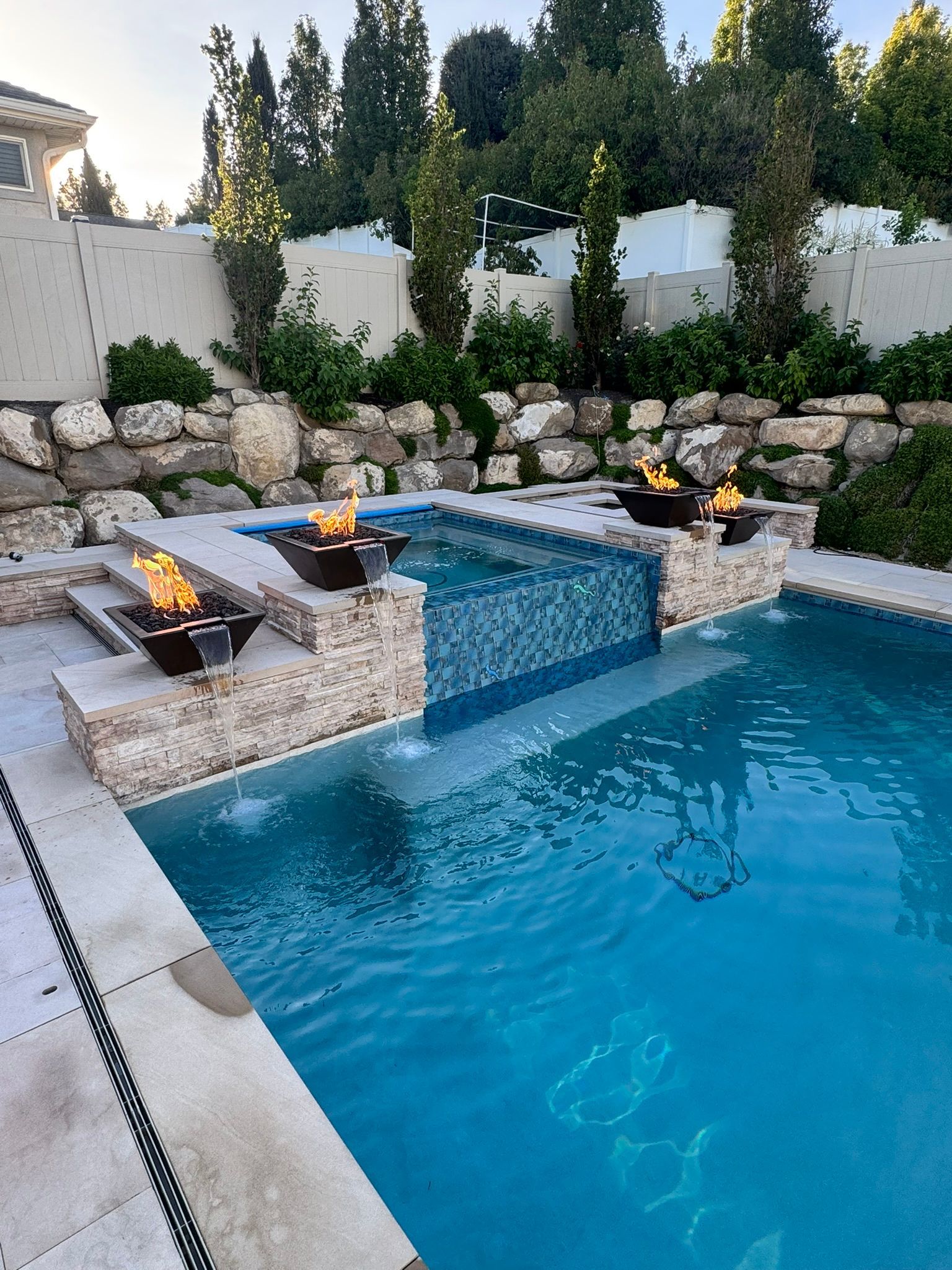 A backyard swimming pool with a raised stone hot tub featuring a tiled spillway, accented by three burning fire bowls.