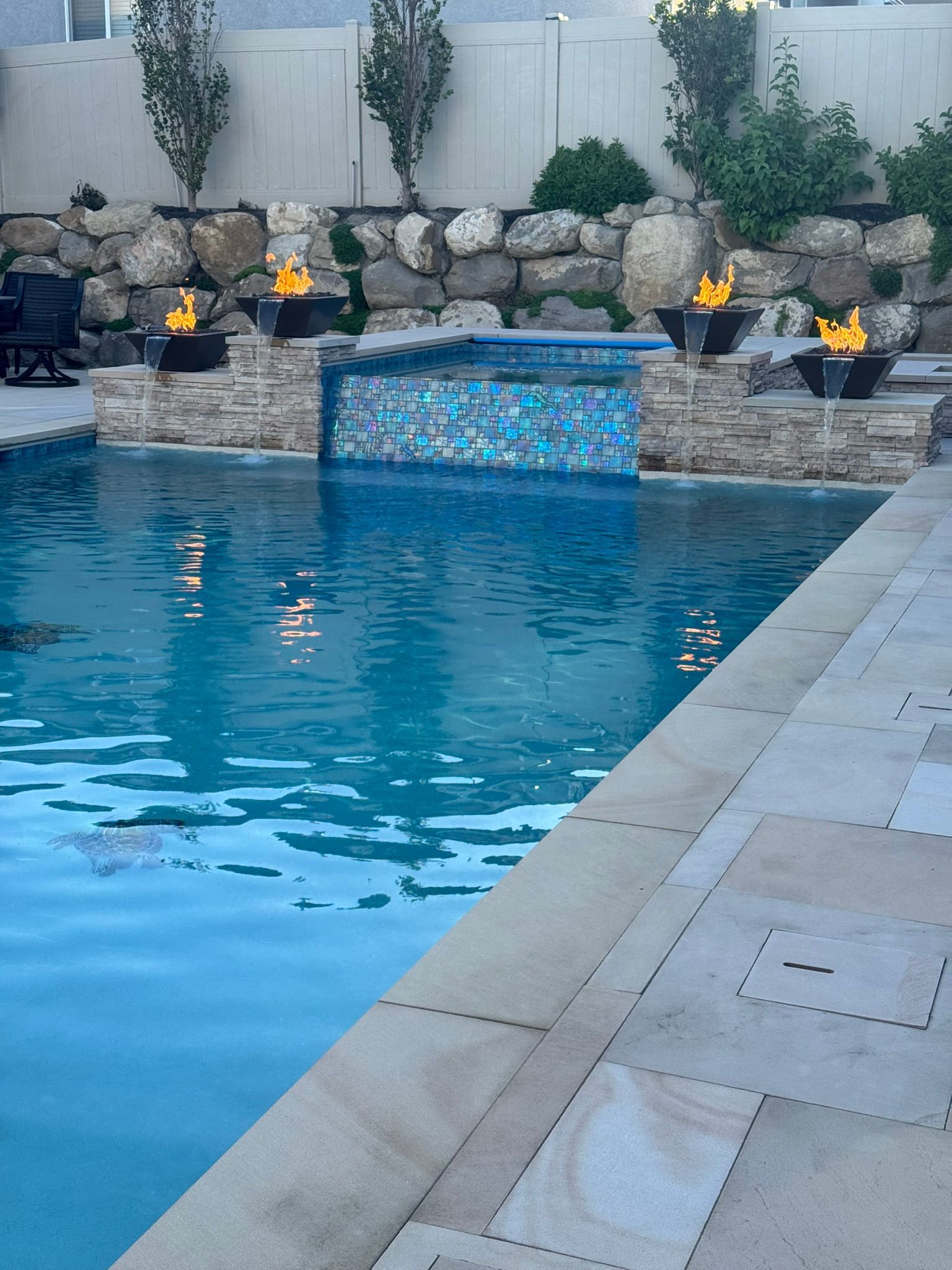 An outdoor pool with four glowing fire bowls, stone accents, and a blue tiled waterfall, set against a rock wall.
