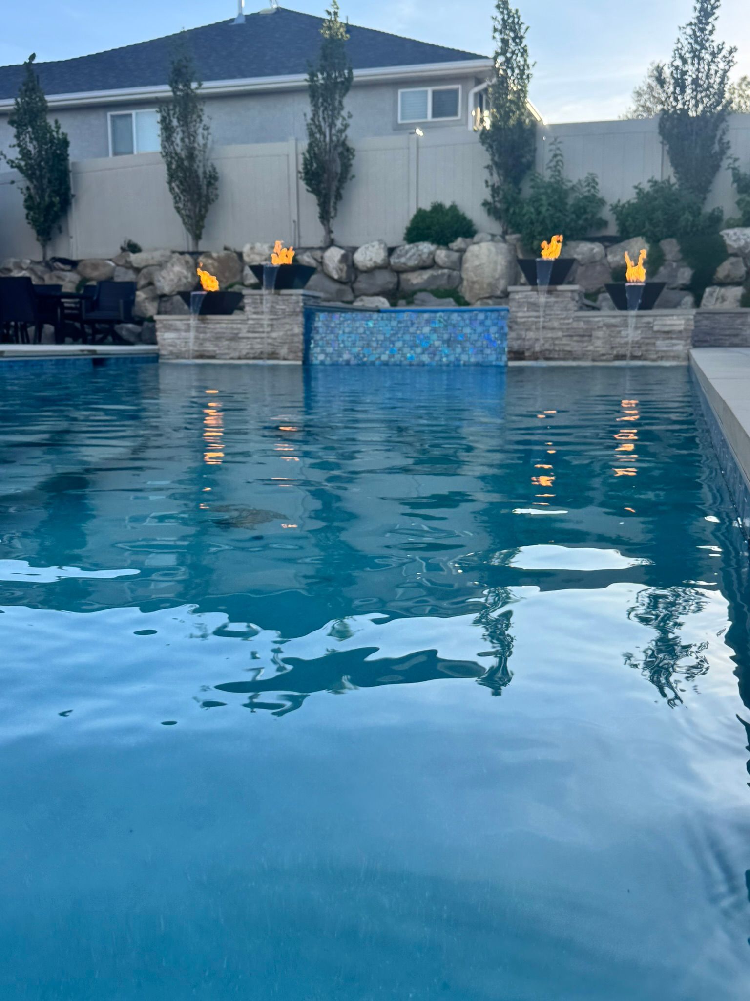A blue backyard swimming pool featuring a decorative tiled water feature wall and glowing fire bowls on stone pillars.