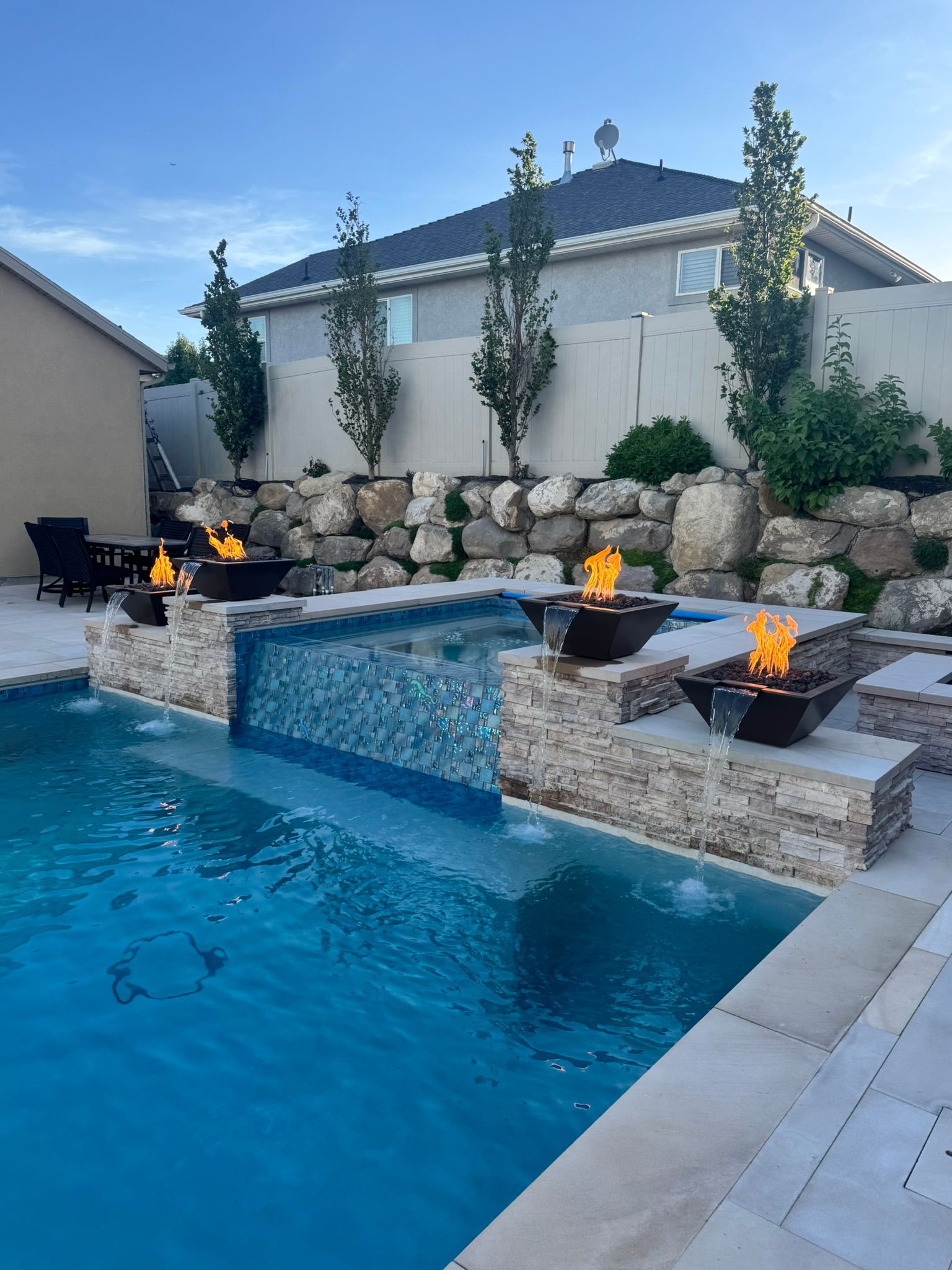 A blue swimming pool with a stone wall, water feature, and three lit fire bowls, set against a house and landscaped hill.
