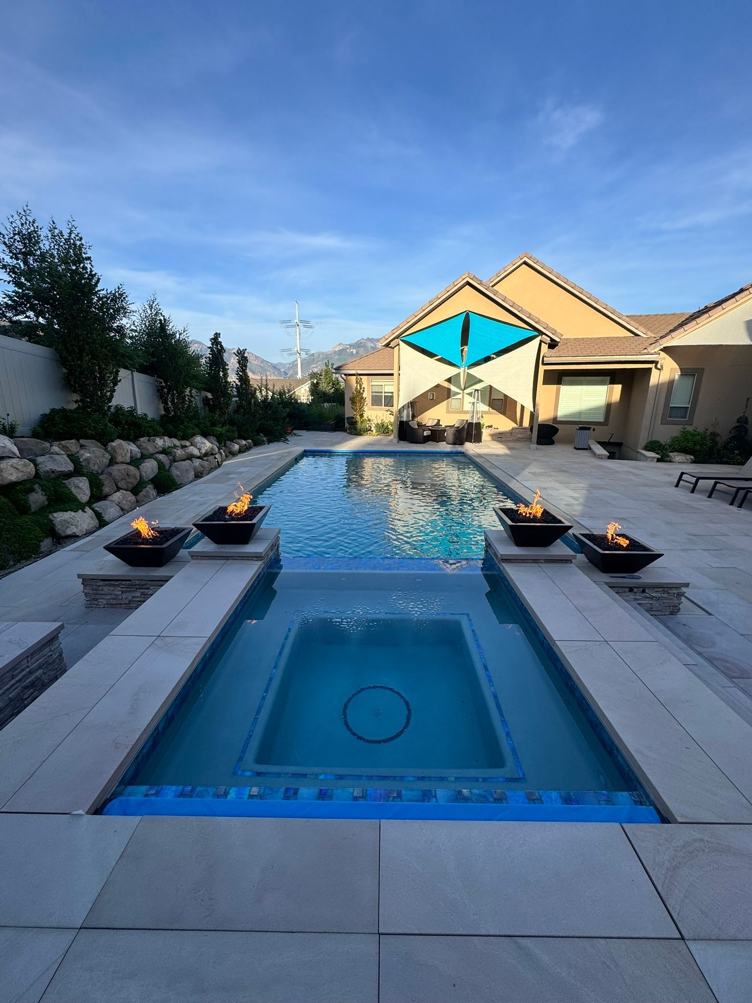 A rectangular swimming pool with an attached spa, flanked by four fire bowls on a paved patio beside a suburban house.