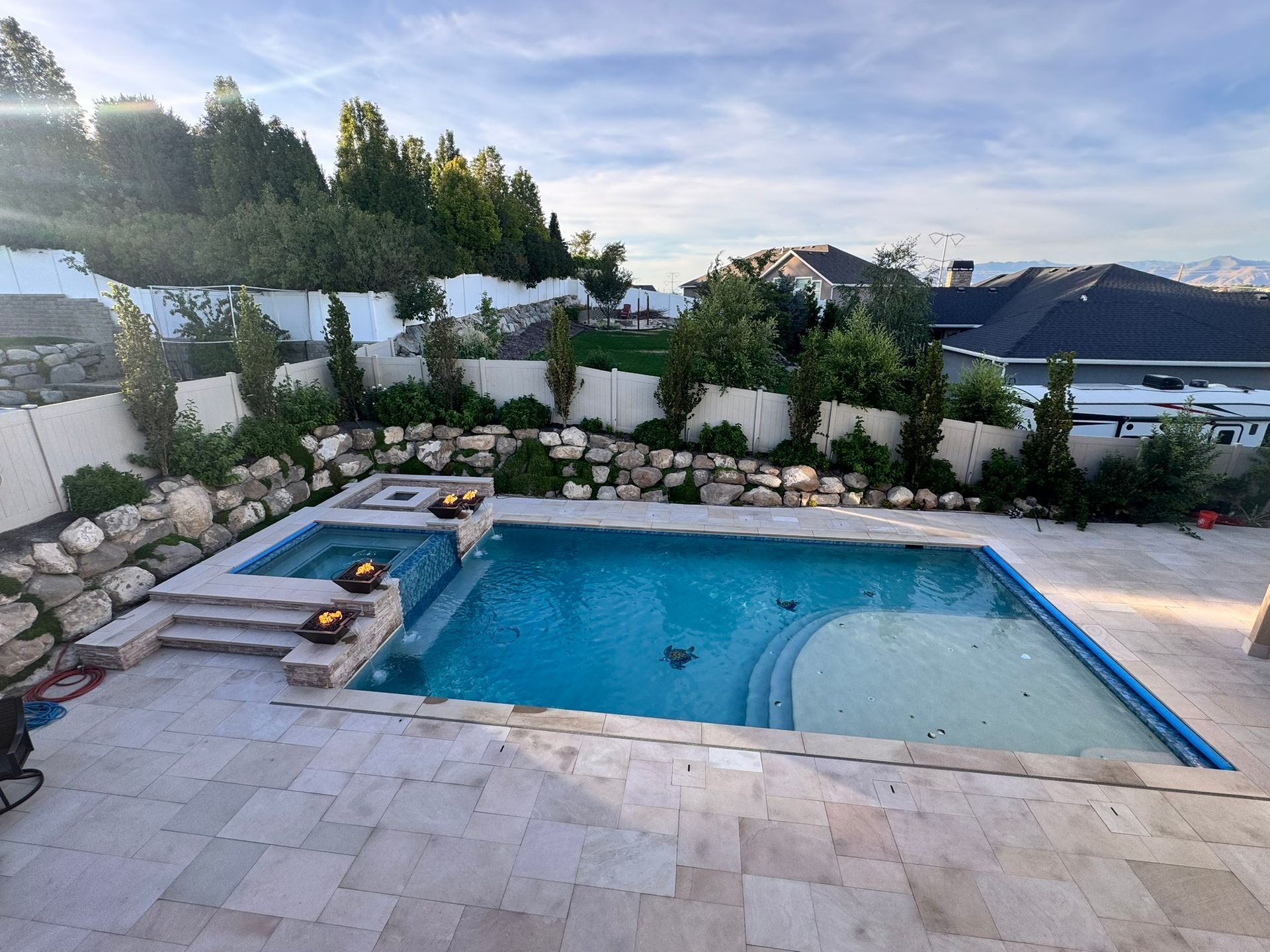 An outdoor swimming pool with an integrated spa, surrounded by stone pavers, a rock retaining wall, and backyard landscaping.