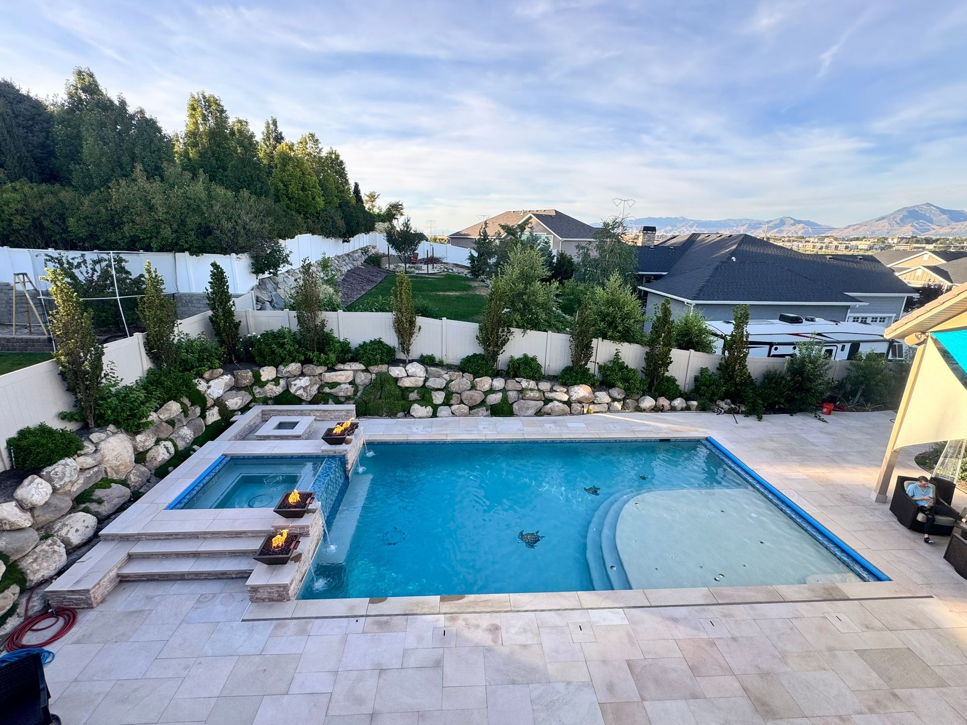 A backyard features a blue swimming pool with an attached spa, surrounded by stone pavers, fire bowls, and a rock wall.