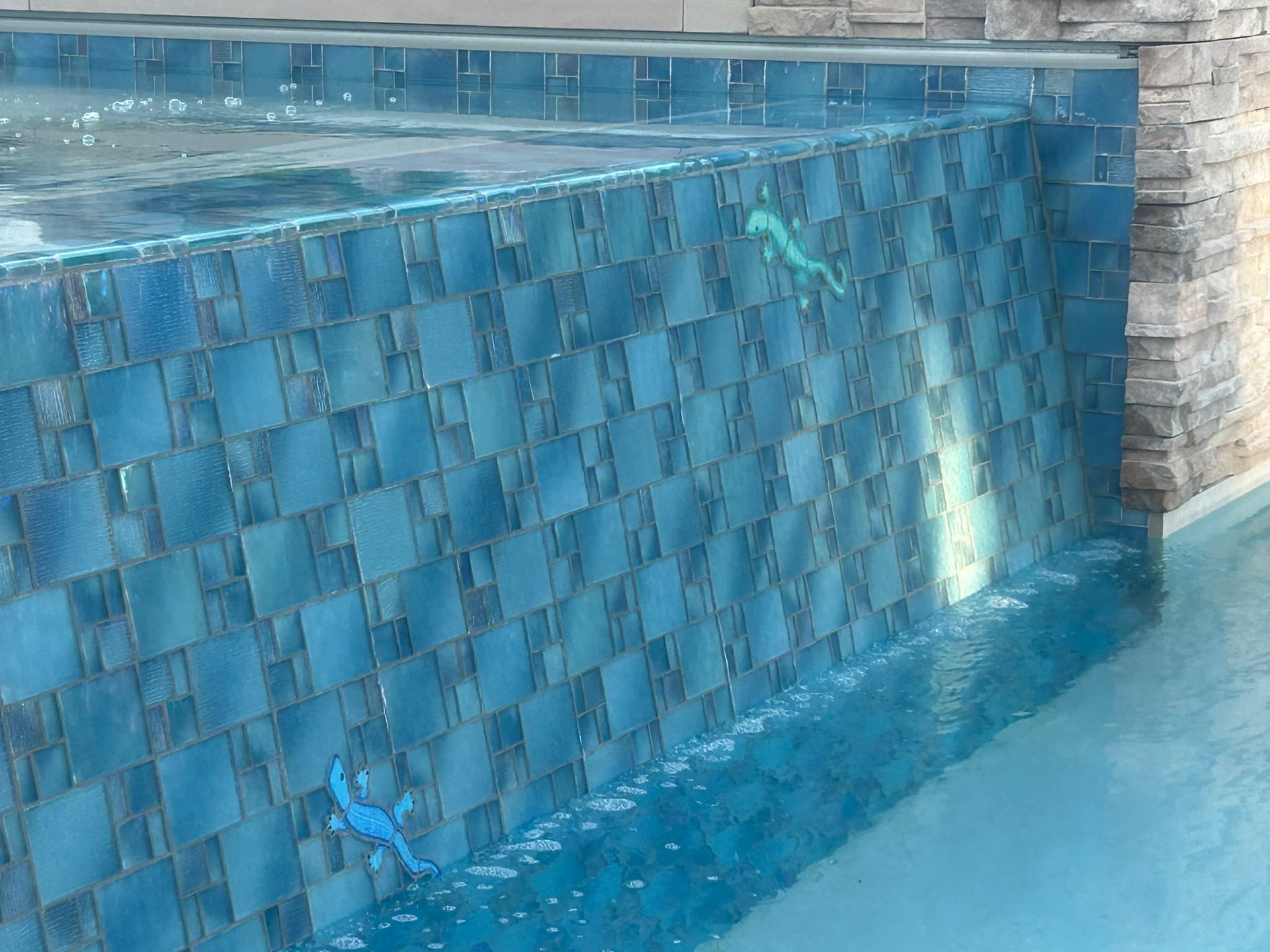Two bright blue lizard decorations are attached to a blue tiled pool wall, with water flowing over the edge.