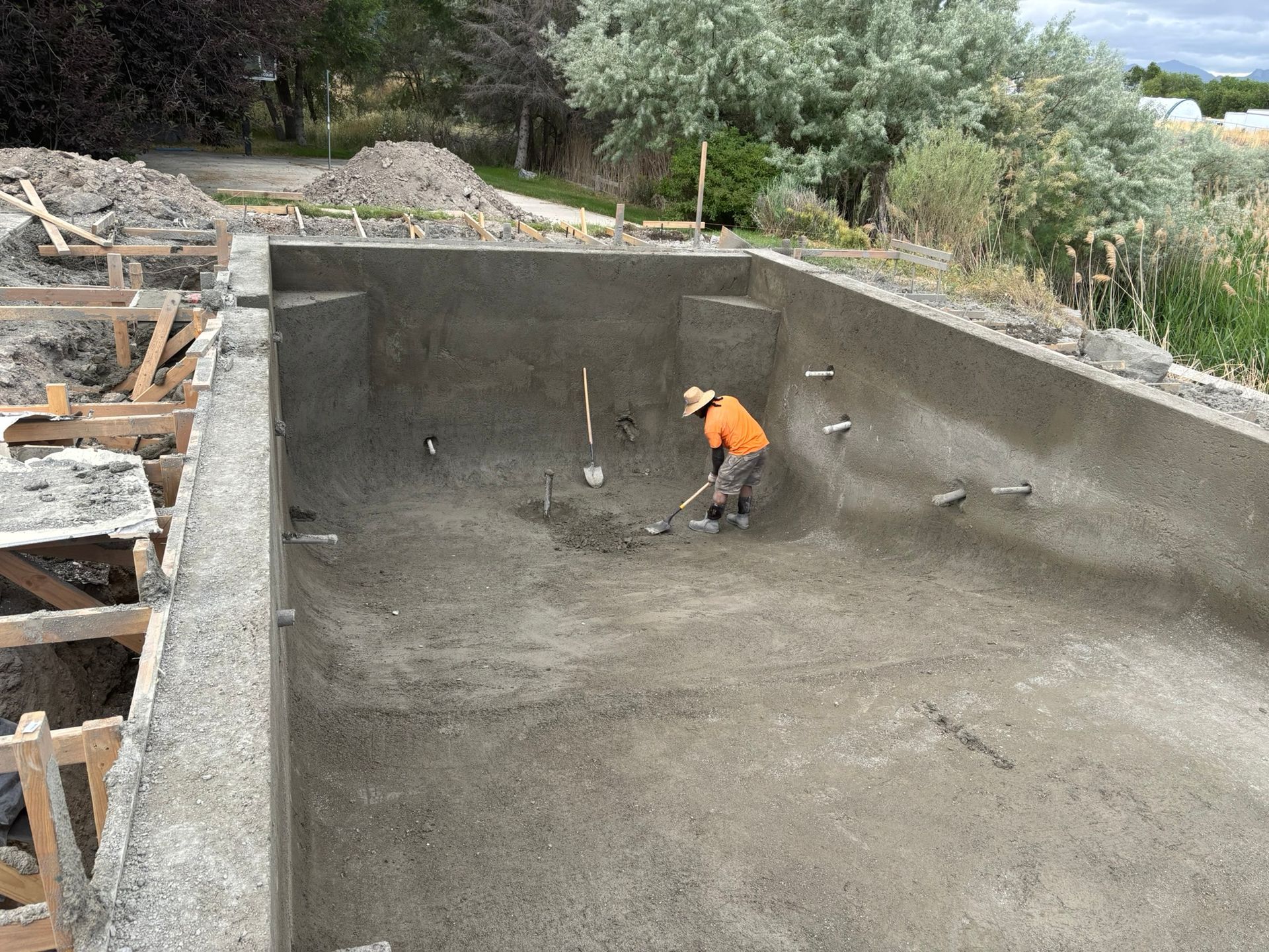 A worker in an orange shirt uses a tool to smooth the concrete floor of a rectangular swimming pool under construction.