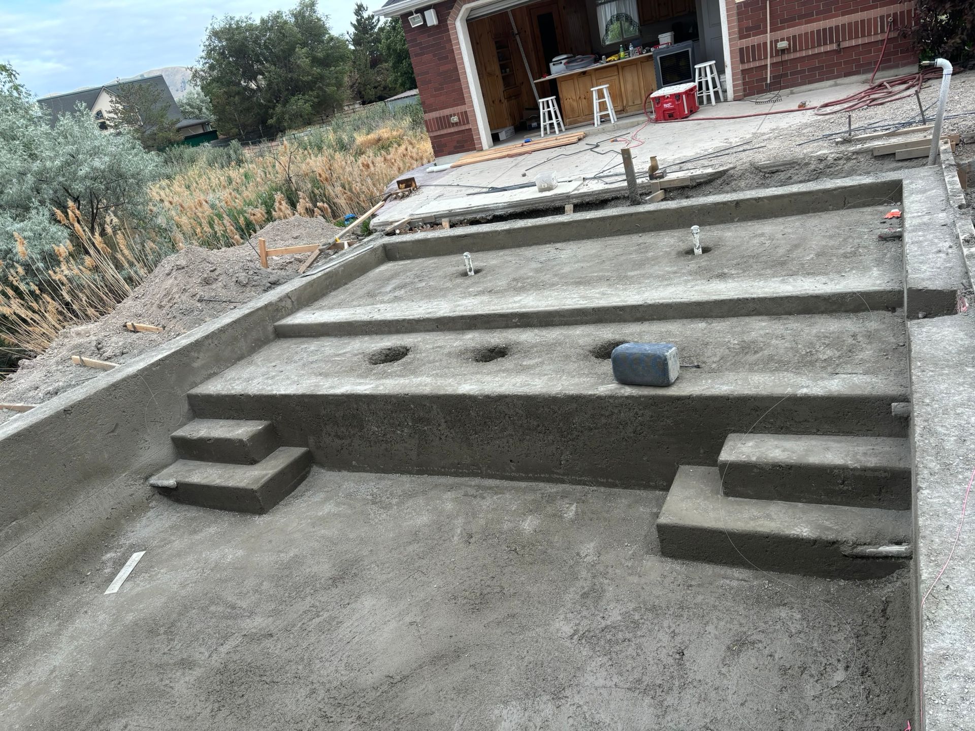 A partially constructed concrete swimming pool with built-in stairs, viewed from above near a house with an open garage.