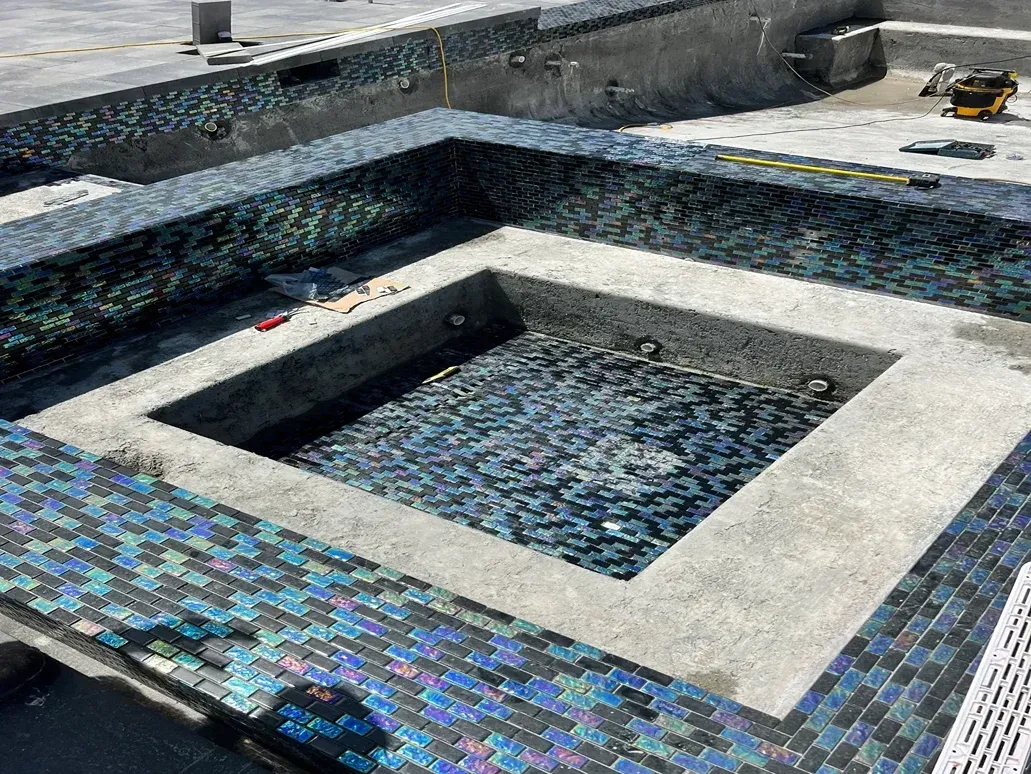 An empty, partially tiled swimming pool or spa under construction, showing blue mosaic tiles and grey concrete surfaces.