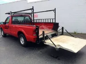 Red pickup truck with a wooden platform ramp at the back and roof racks parked outside.