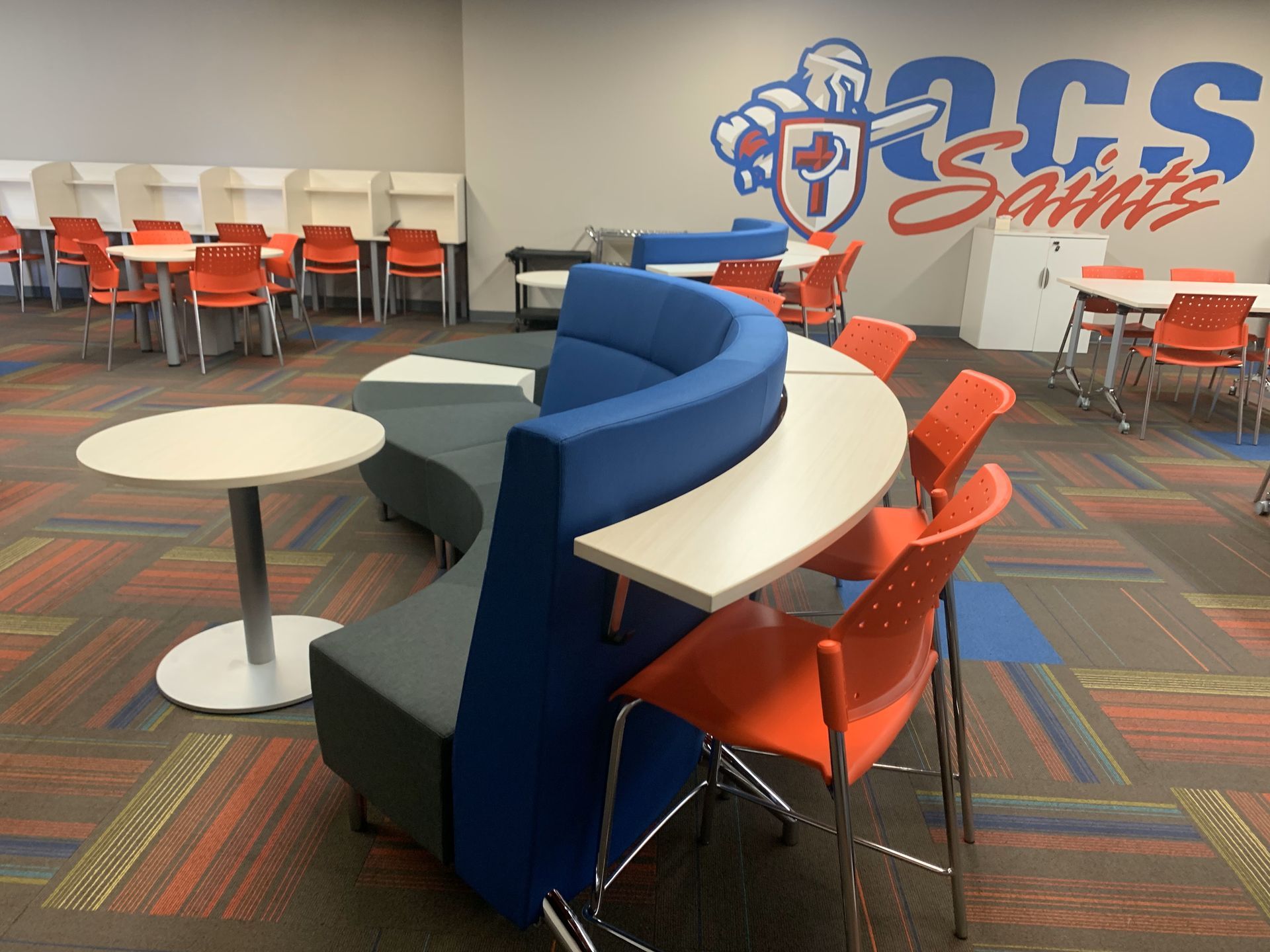 Side view of blue curved high back modular sofa with white work counter and red stools; white tables and red chairs in back.