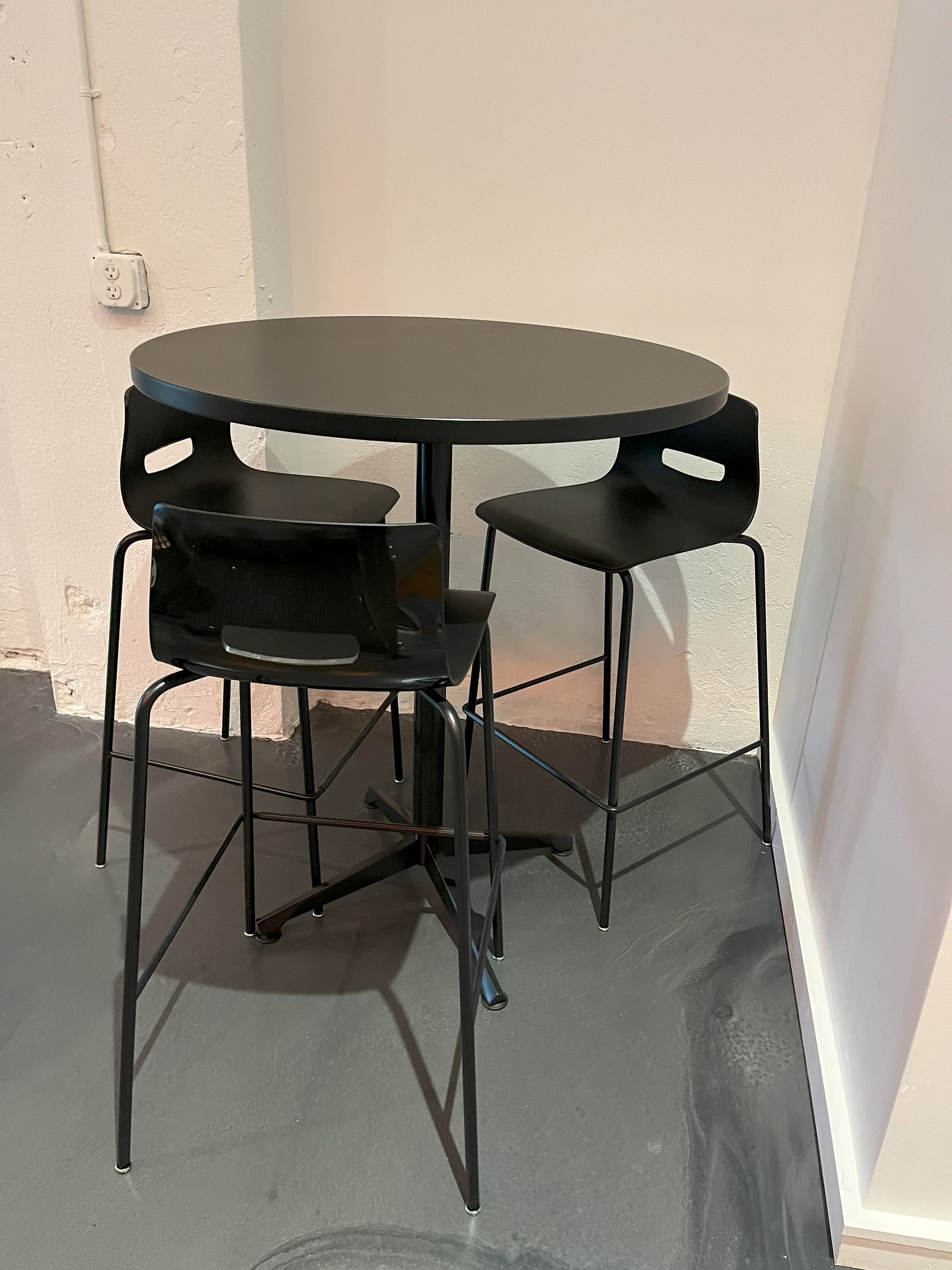 Round black bar table with 3 black stools in a corner, against white walls.