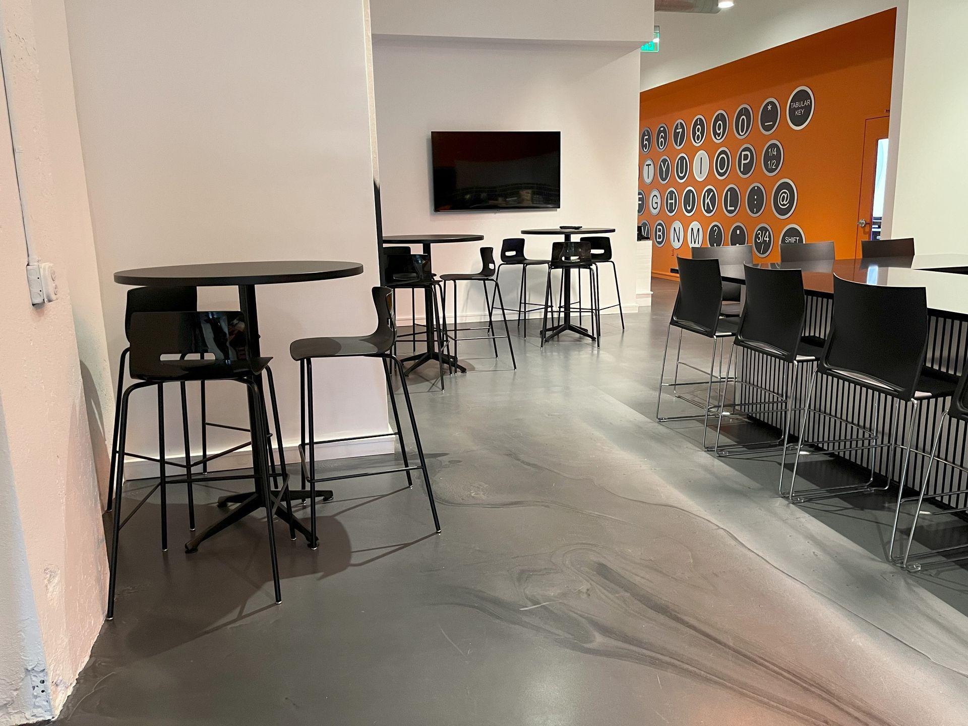 Breakroom with 3 round black cafe tables and black stools, black counter stools; concrete floor with orange wall behind.
