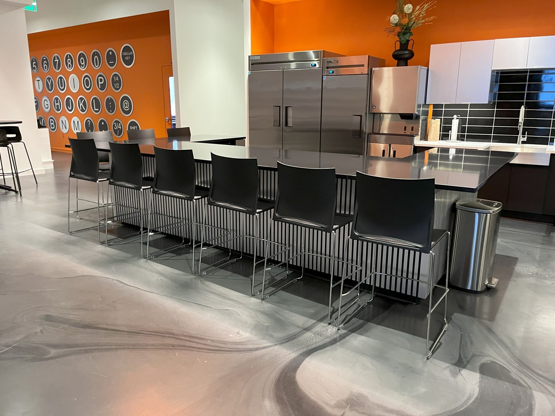 Breakroom kitchen with black stools at a black and white counter, stainless steel appliances and orange wall behind. 
