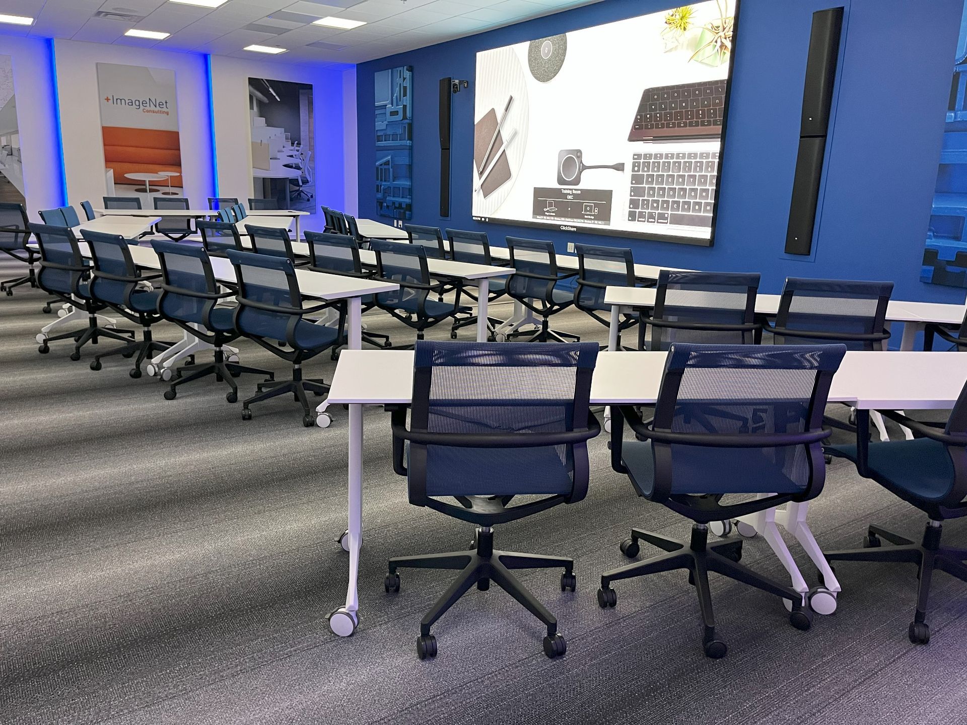 A classroom with tables and chairs and a large screen on the wall.