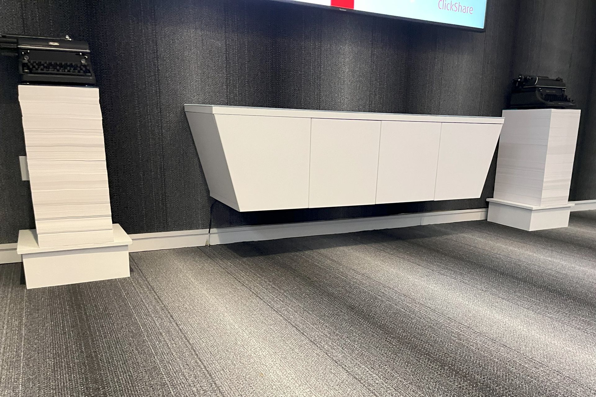 Angled floating credenza on gray striped wall; white pedestals with typewriters on either side. 