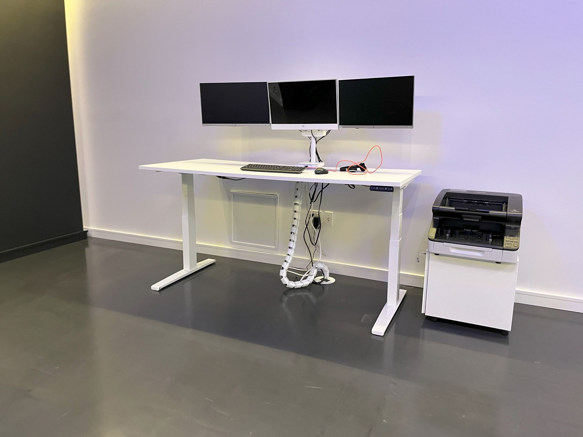 All white height-adjustable desk with 3 monitors and white cable management spine on concrete floor.