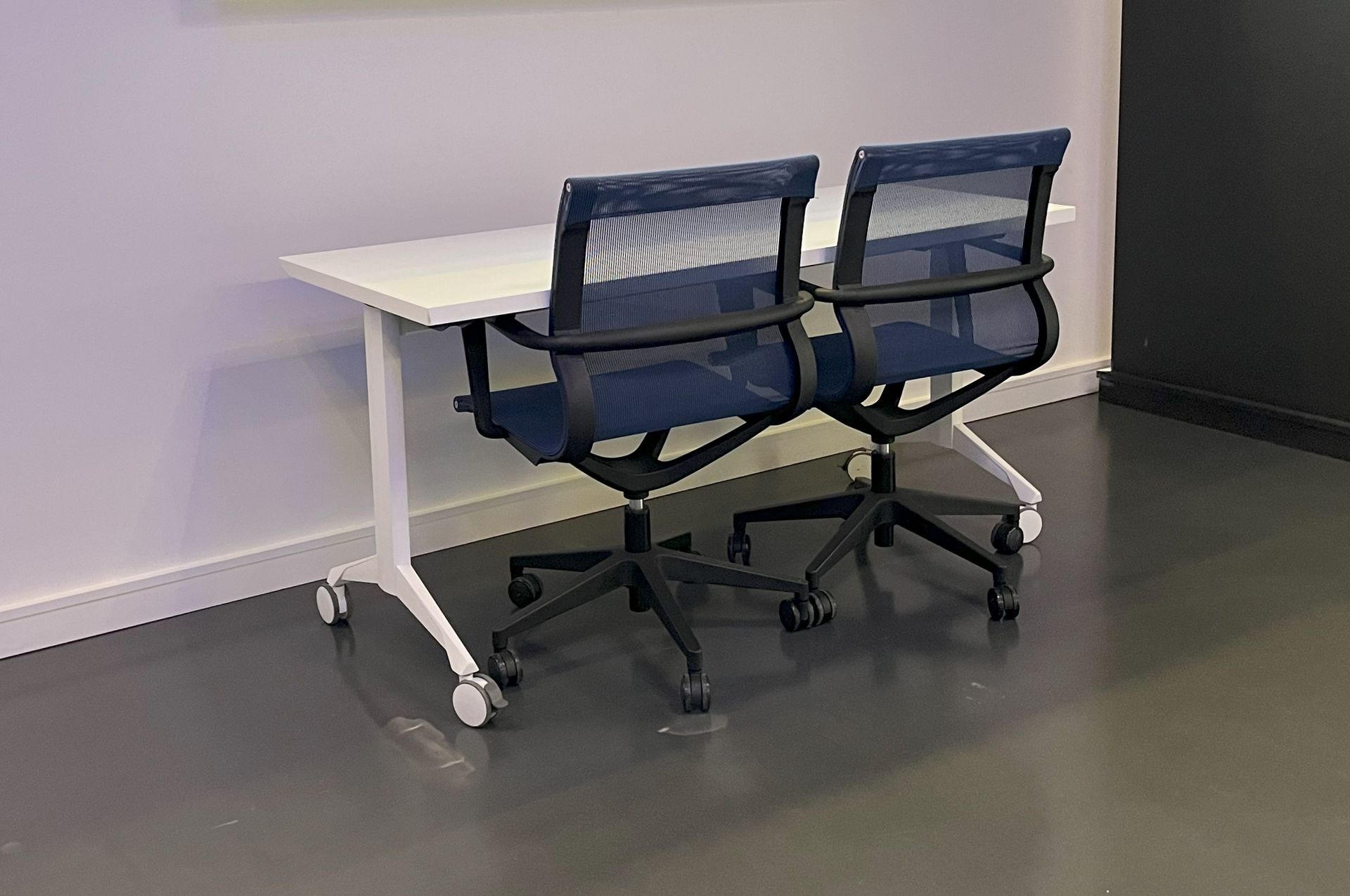 All white training table with two blue mesh chairs against a white wall and concrete floor.