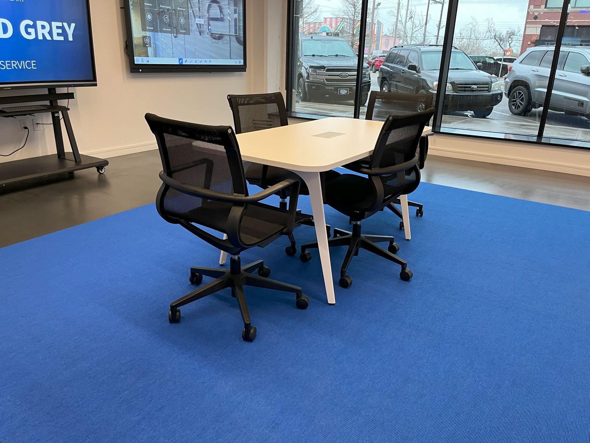 Small white rectangular meeting table with 4 black chairs on blue rug with windows in background.