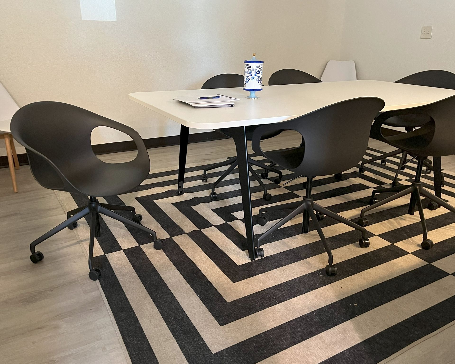 A small white modern conference table with black plastic bucket chairs on a striped rug.