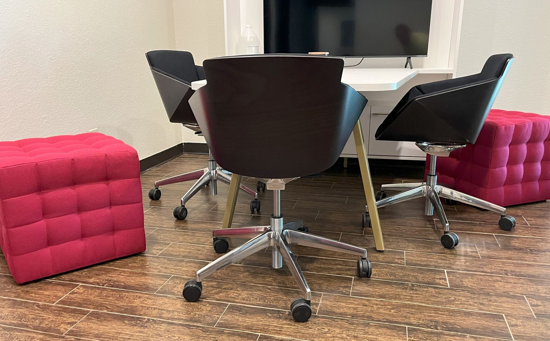 Meeting space with white AV table & large TV, black wood shell conference chairs, and pink tufted cube ottomans.