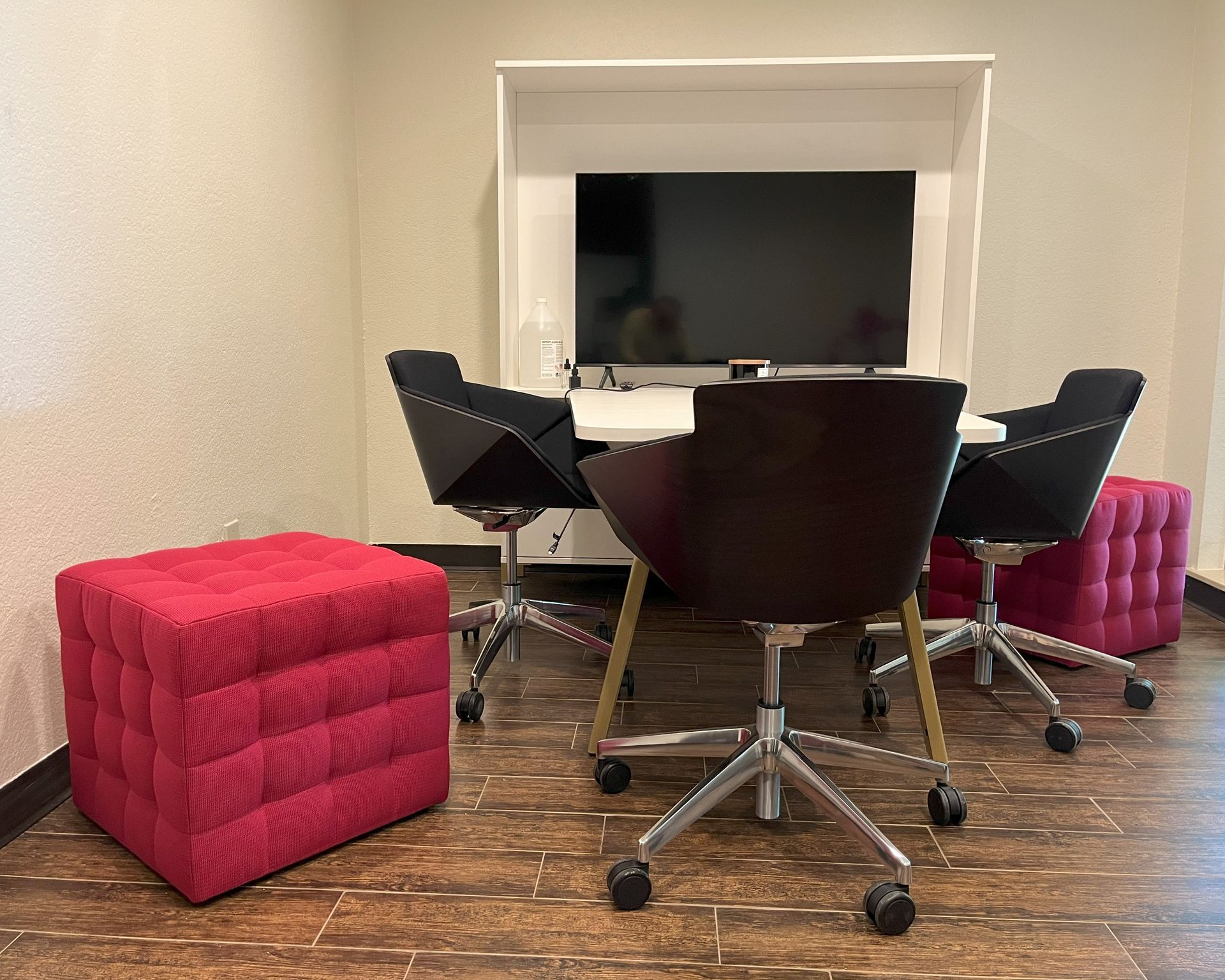 A room with a white AV meeting table, black rolling chairs, pink cube ottomans and a flat screen tv.