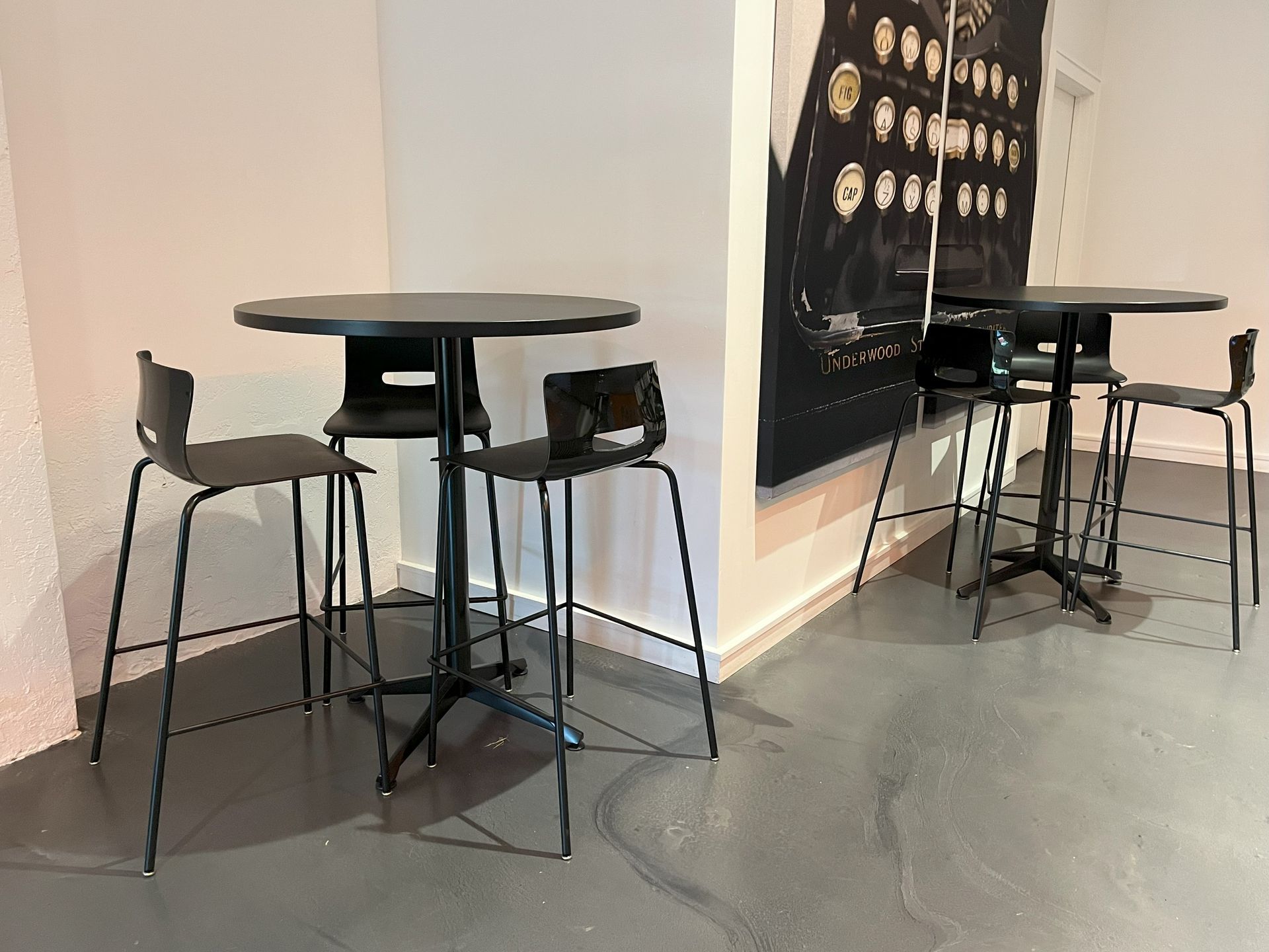 Two round black bar tables each with 3 black stools; large canvas print of a typewriter behind back table.