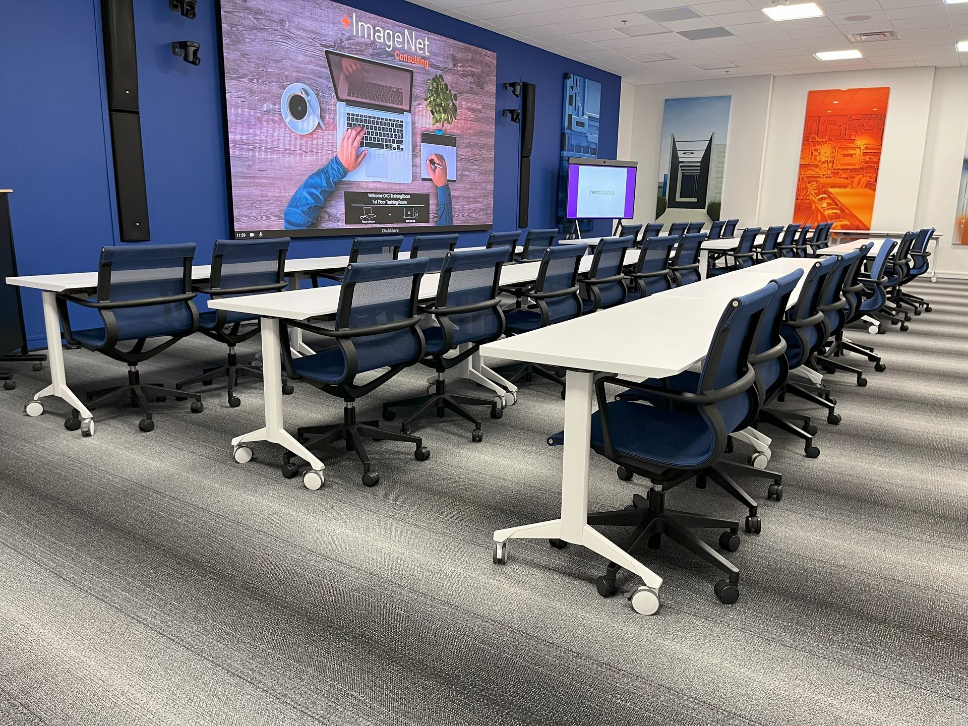 Three rows of all white training tables with blue mesh chairs in a room with gray striped carpet and a blue wall.