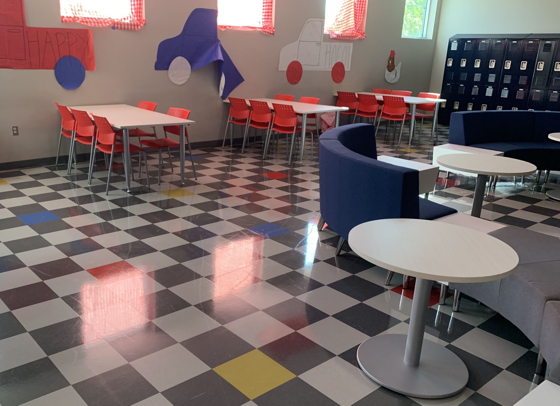 Large room with white tables and red chairs, lounge seating and a checkered floor; blue lockers along back wall.