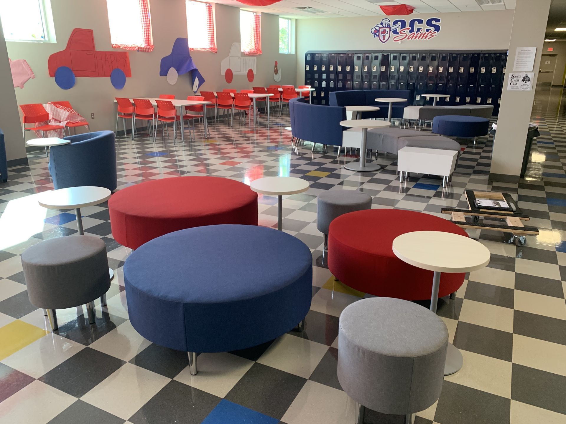 Large room with checkered floor and lockers; blue, red, and gray round ottomans with tables in front, various seating behind.