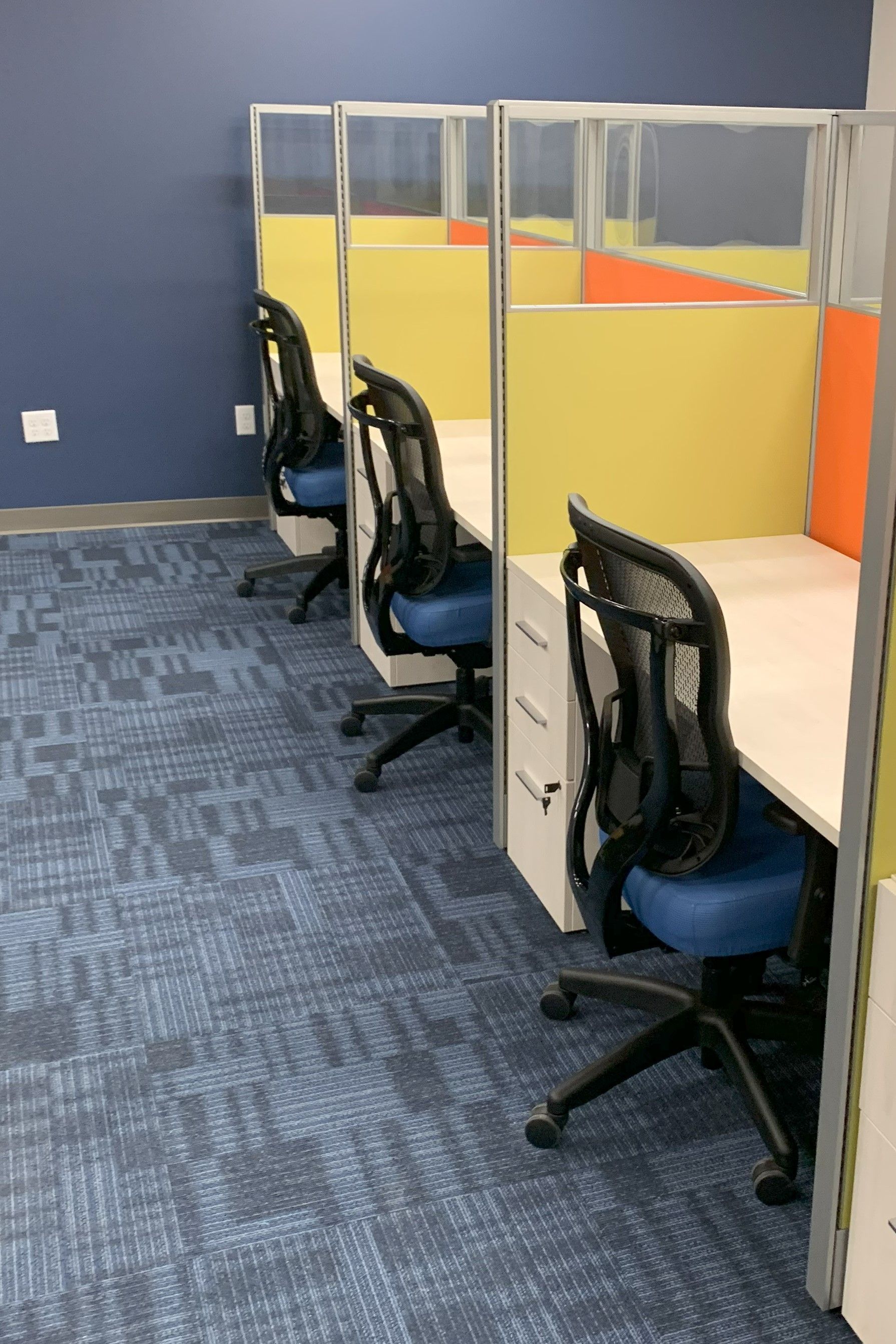 Office cubicles with light green and orange panels, black mesh chairs with blue seat, and blue carpet.