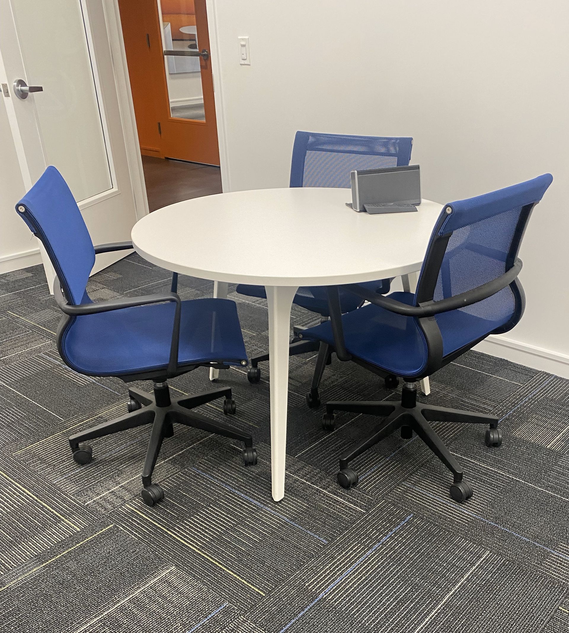 Room with white round table and 3 blue mesh chairs; white walls and striped gray carpet.