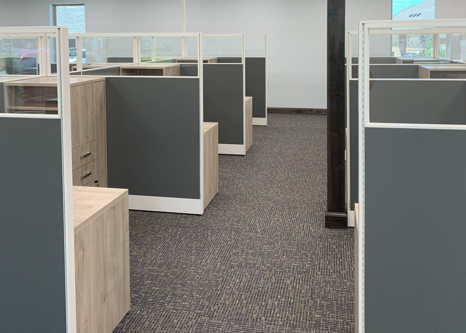 Office cubicles with gray and wood-toned furniture. Clear glass dividers and neutral carpet.