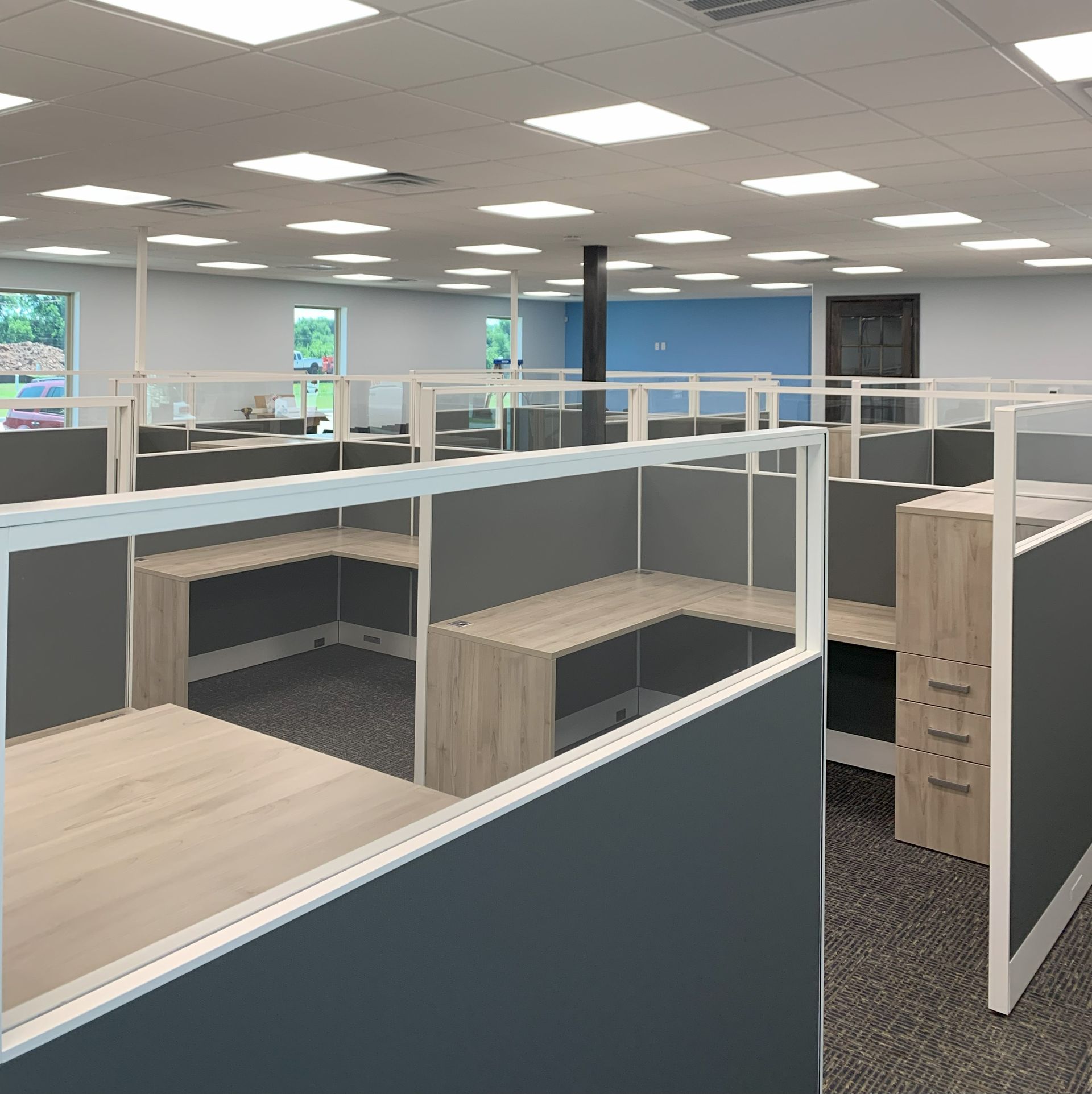 Office cubicles with gray and tan desks and gray panels, under bright white ceiling lights.