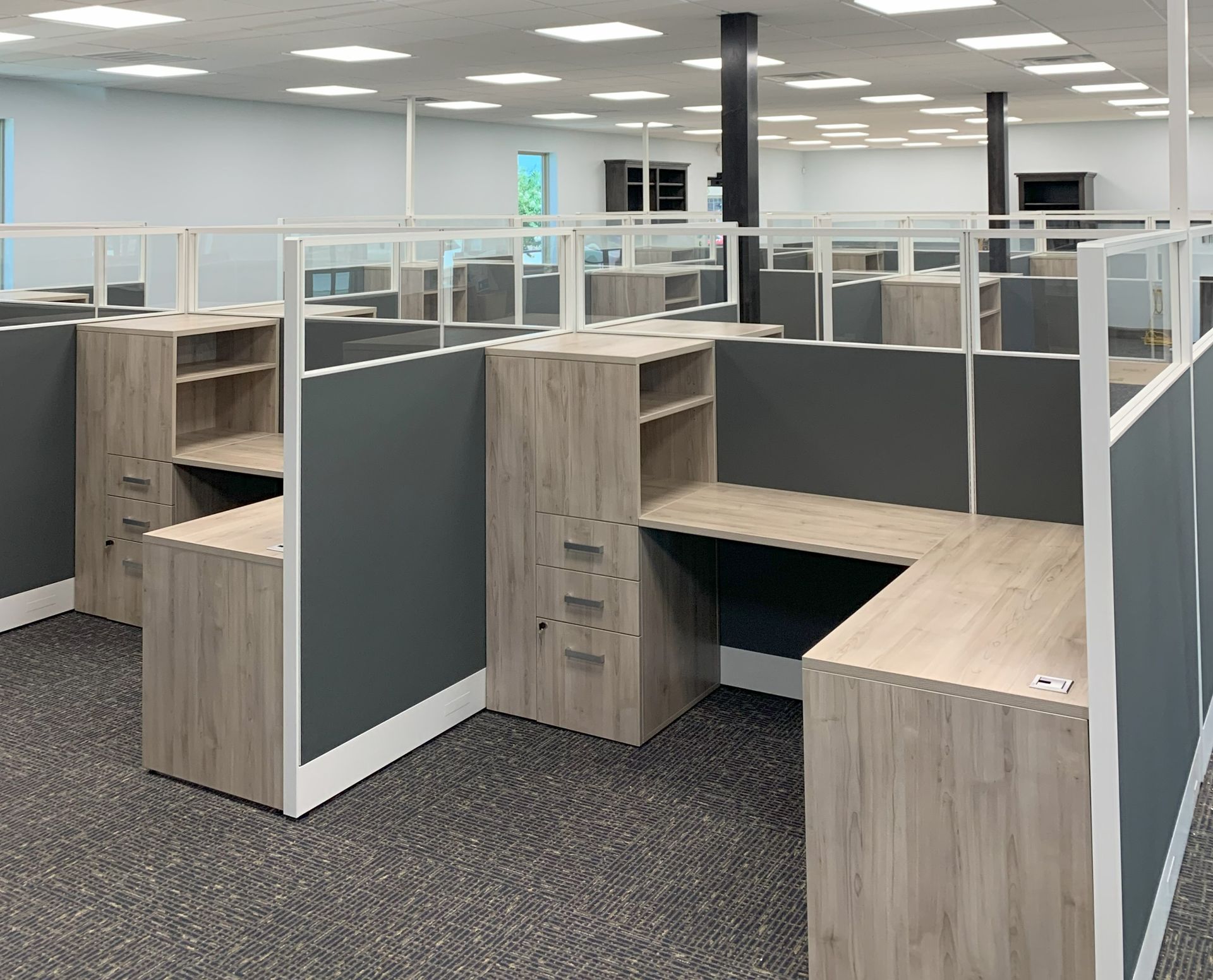 Office cubicles with gray panels, light wood desks, and clear glass dividers in a large, bright room.