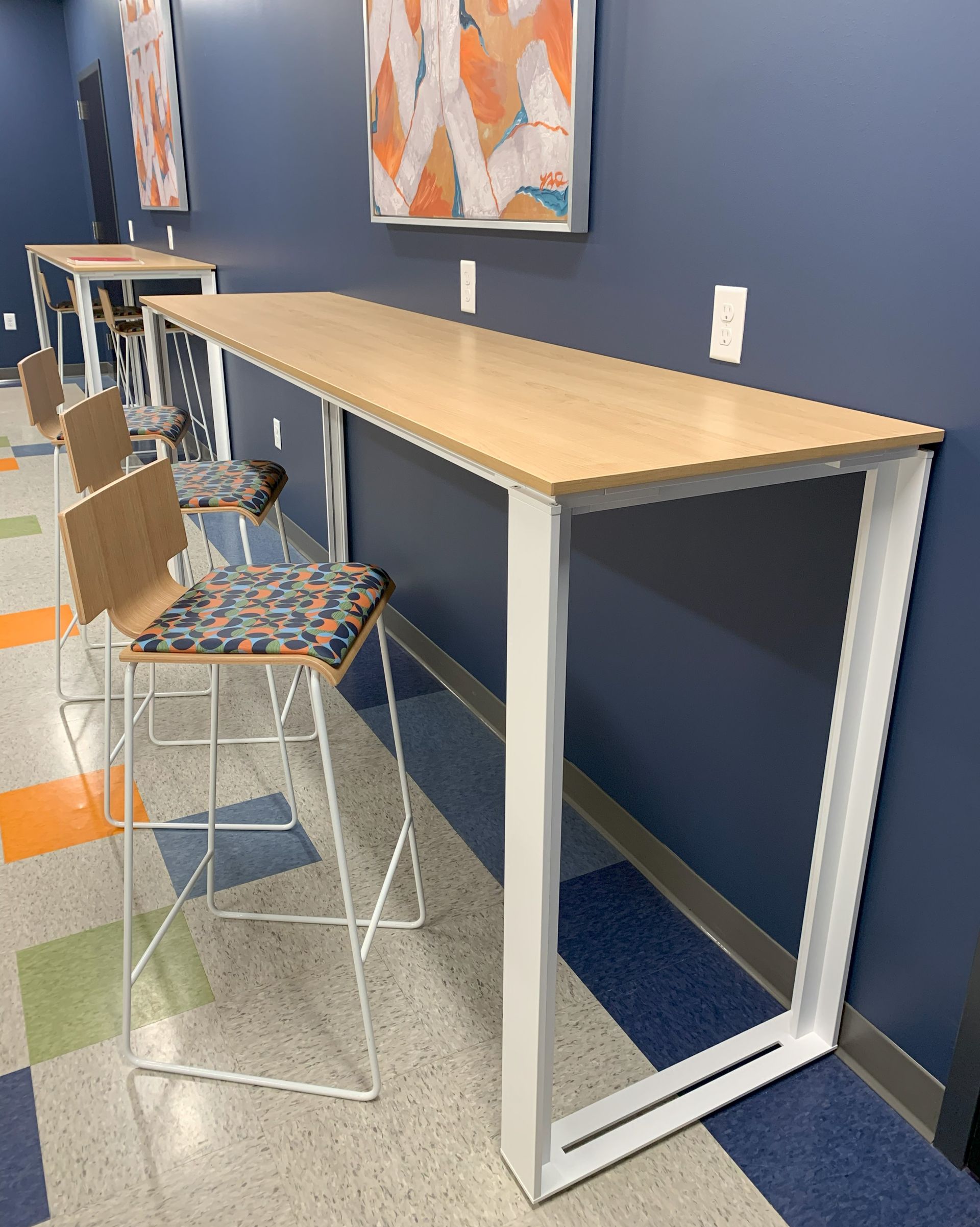 Long, light wood table with white metal legs and patterned stools against a blue wall, artwork above.