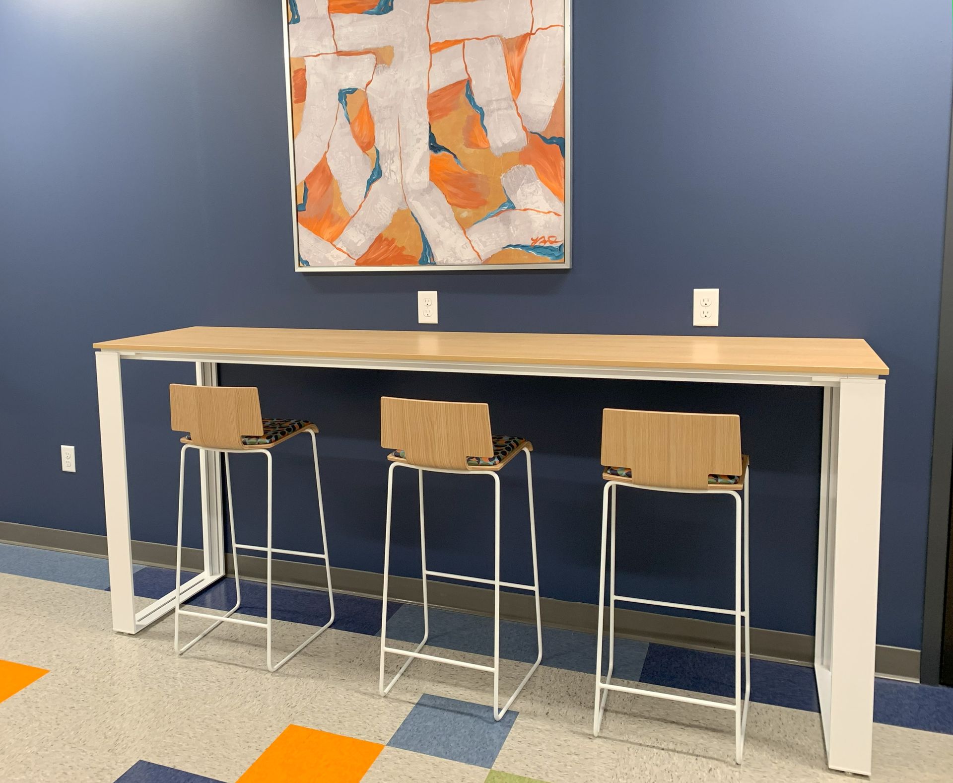 Three bar stools at a long cafe table against a blue wall with abstract art; orange and blue floor.