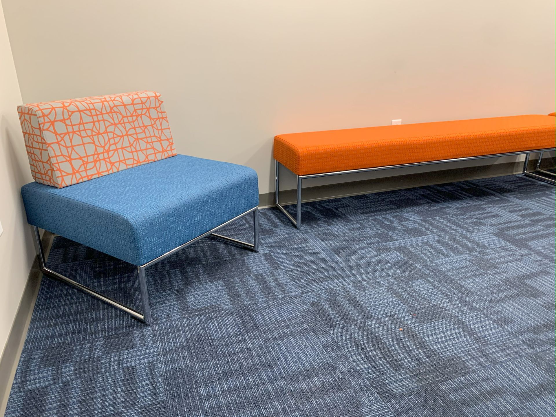 Upholstered chair with blue seat and orange patterned back next to long orange bench, against a corner wall on blue carpet.