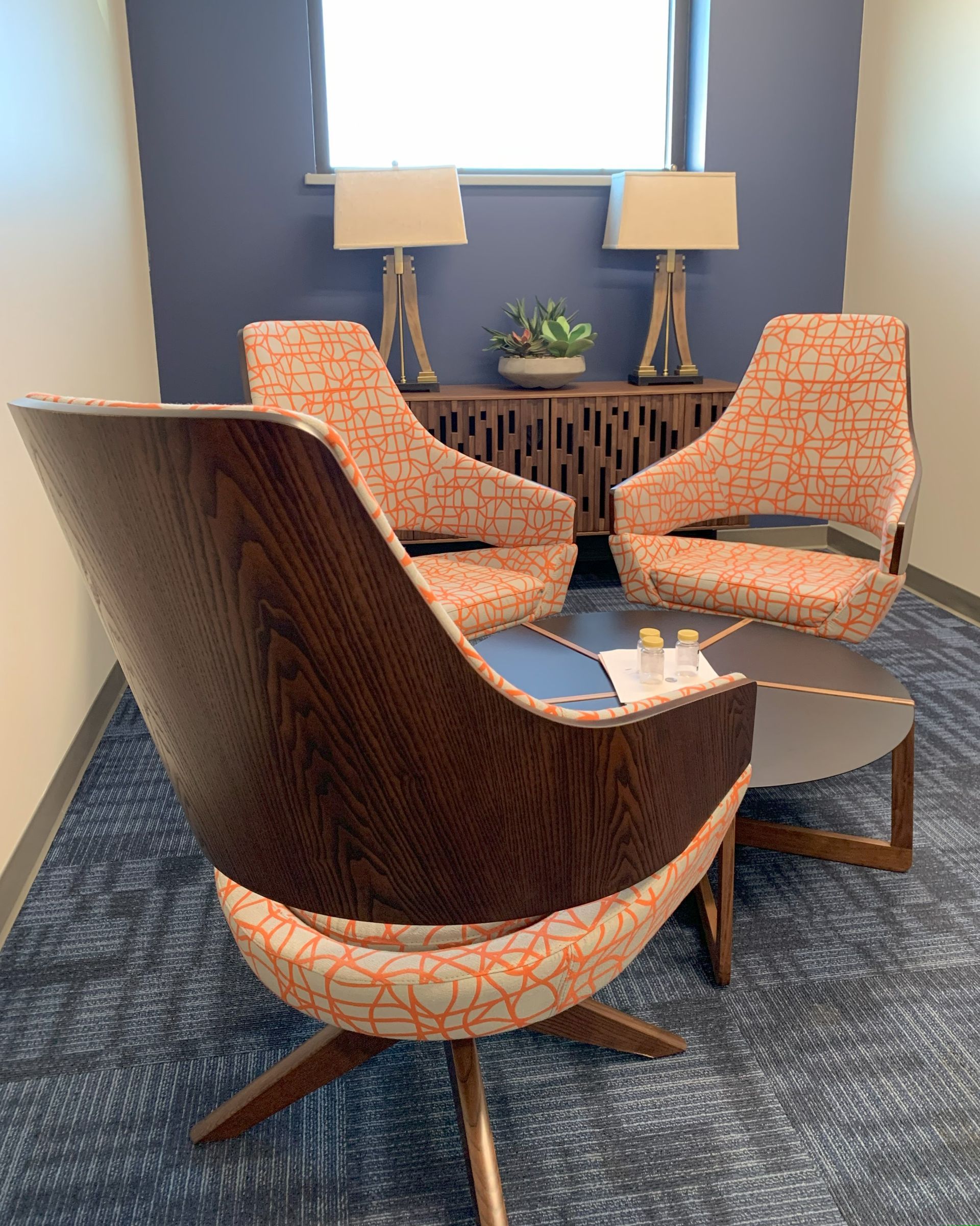 Three orange patterned chairs with wood frame around a circular table in a blue-carpeted room, in front of cabinet with two lamps and a dark blue wall.