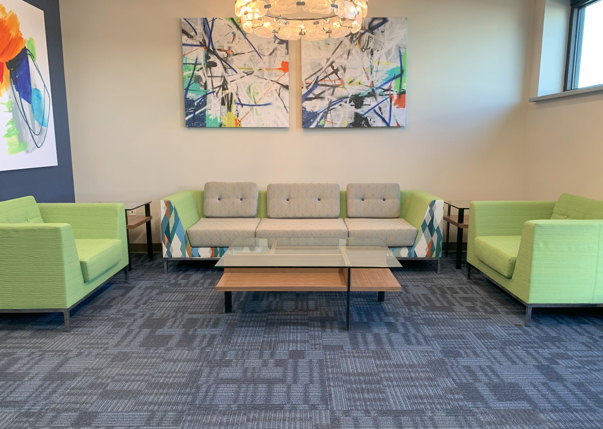 Lounge area with two green armchairs facing each other on either side of a long colorful sofa in multiple patterns, and a coffee table in the middle. Paintings hang on the wall.