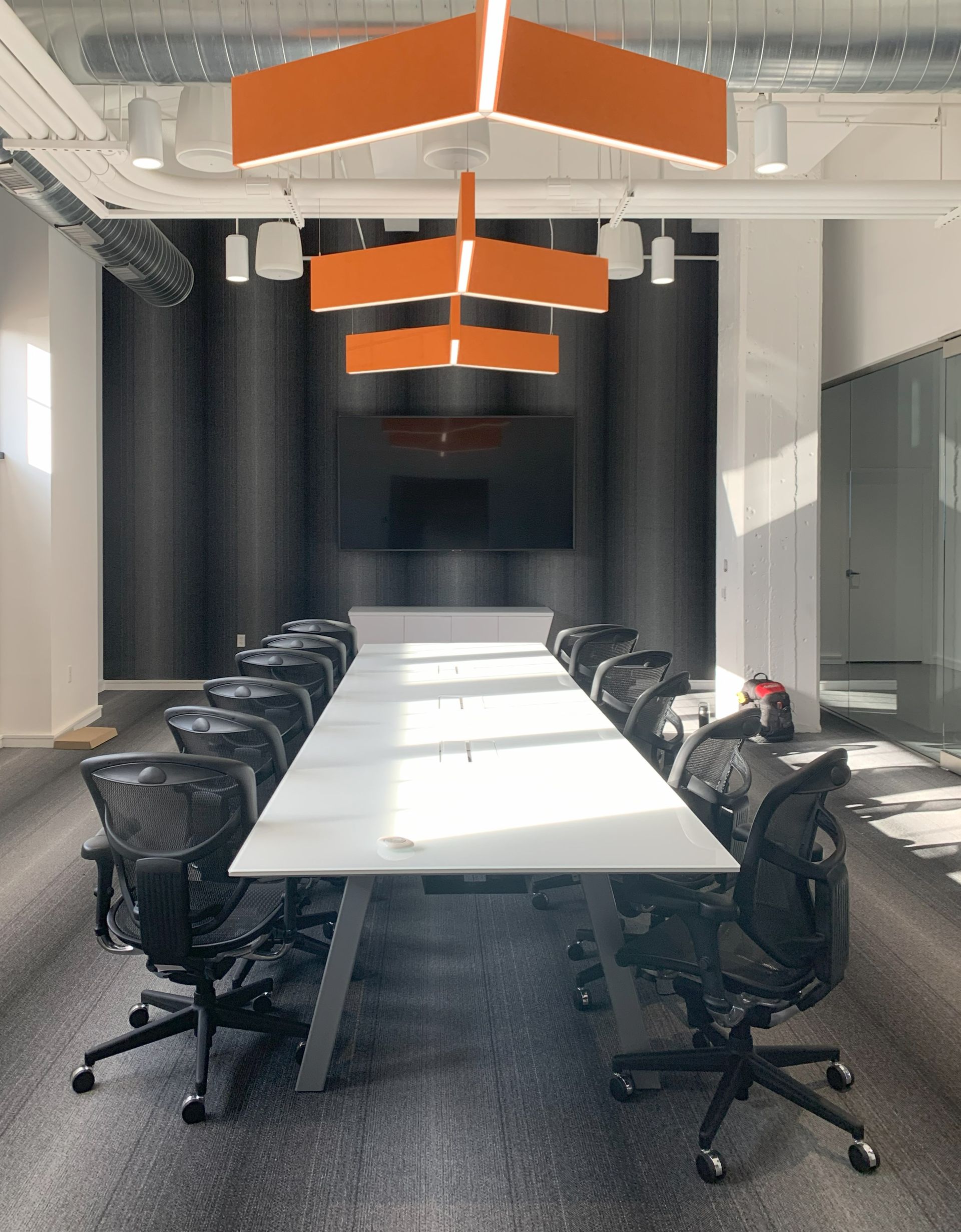 End view of white rectangular boardroom table surrounded by black chairs in front of TV on dark gray wall; orange light above