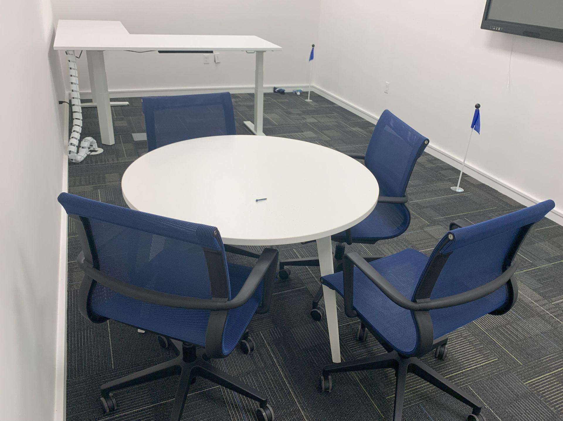 Office with white walls and gray carpet, round white meeting table with 4 blue mesh chairs in front of white desk.