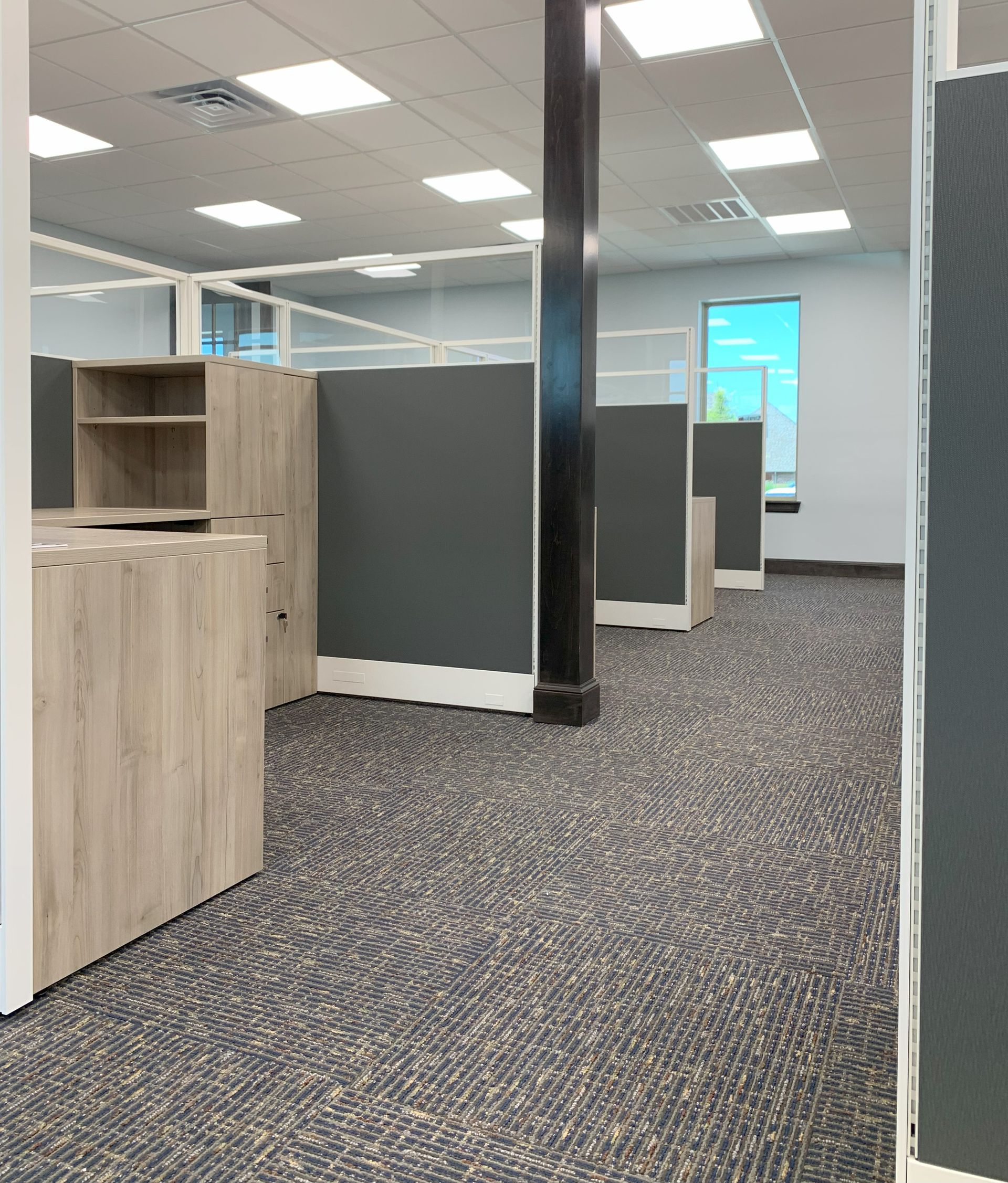 Office cubicles with gray and wood-grain desks, dark carpet, and overhead lighting.